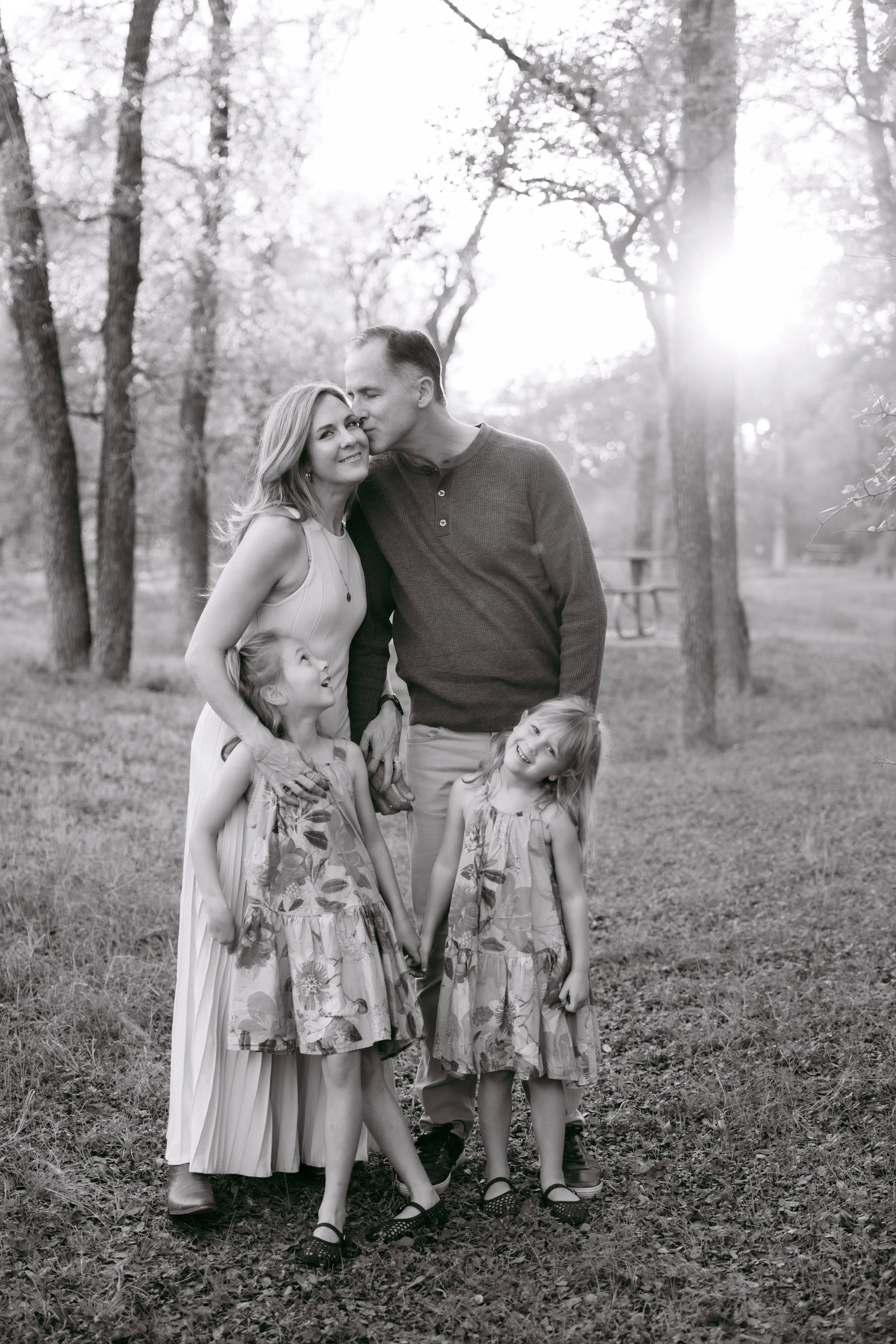 A family poses outdoors in a wooded area, with a man kissing a woman on the forehead and two young girls smiling and holding hands in front of them, during sunset or sunrise.