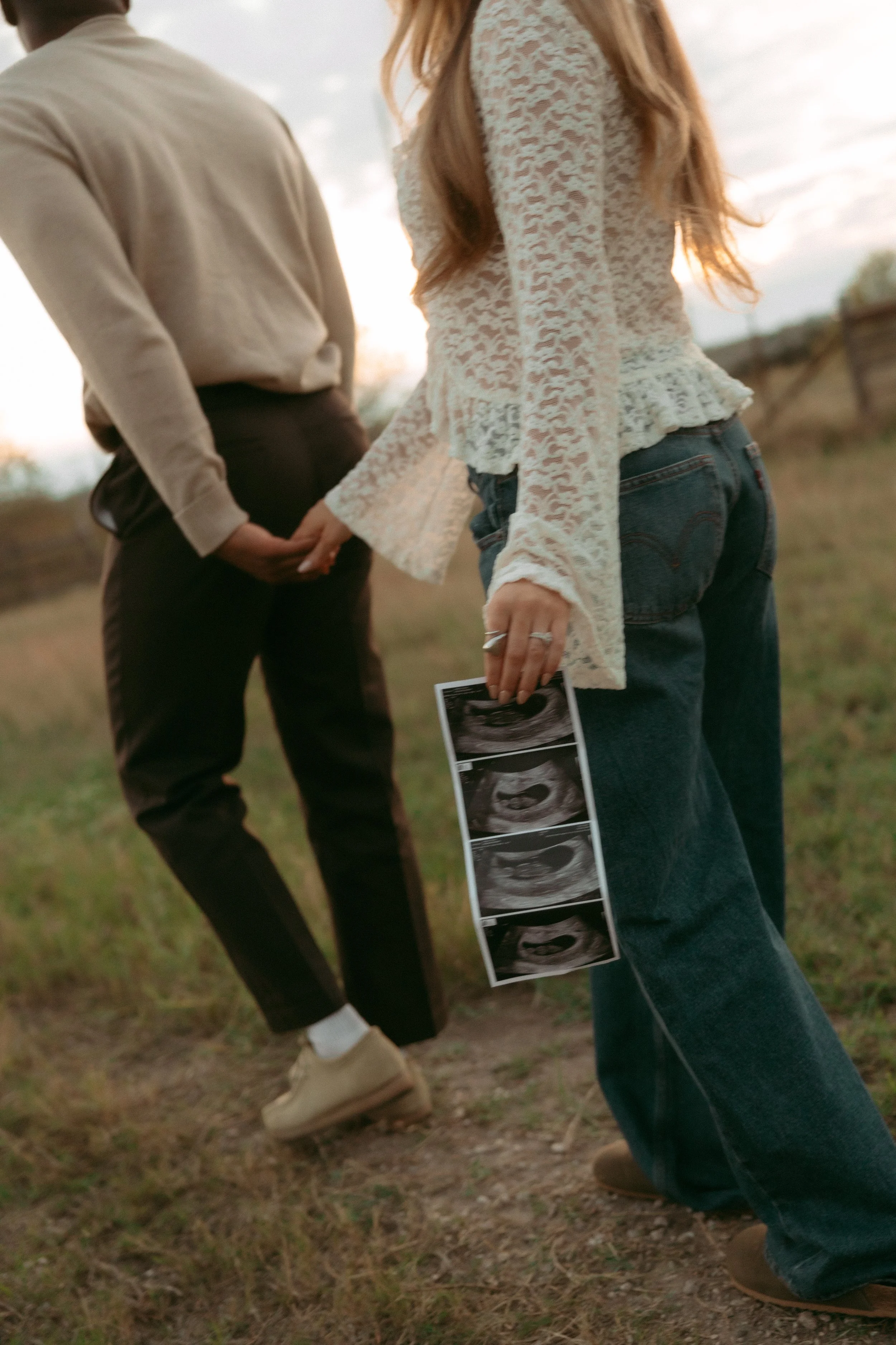 A woman holding ultrasound pictures, standing outdoors with a man in the background, both in casual clothing, during sunset.