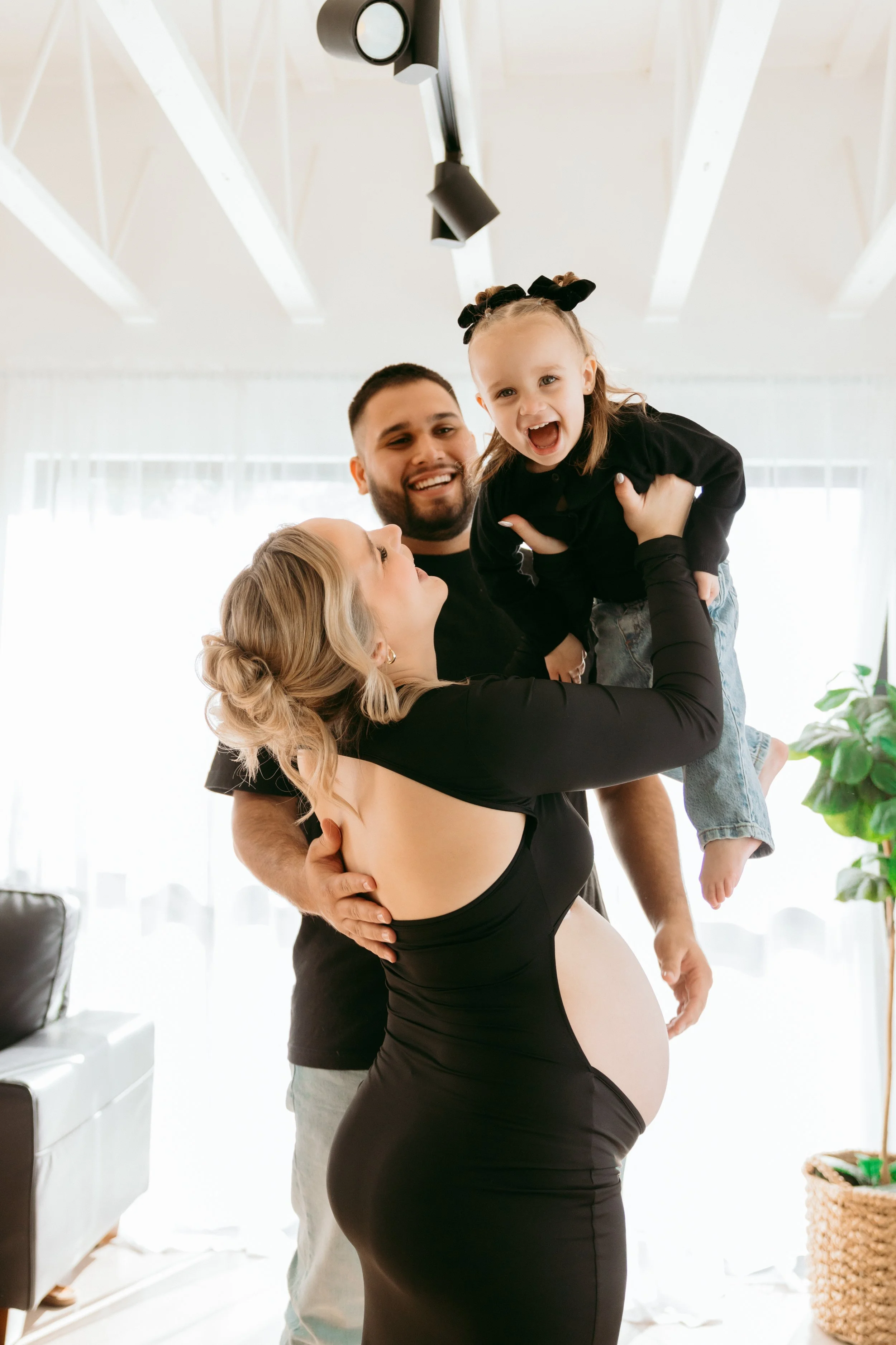 Family celebrating pregnancy in living room with a pregnant woman, her husband, and young daughter, all smiling and joyful.