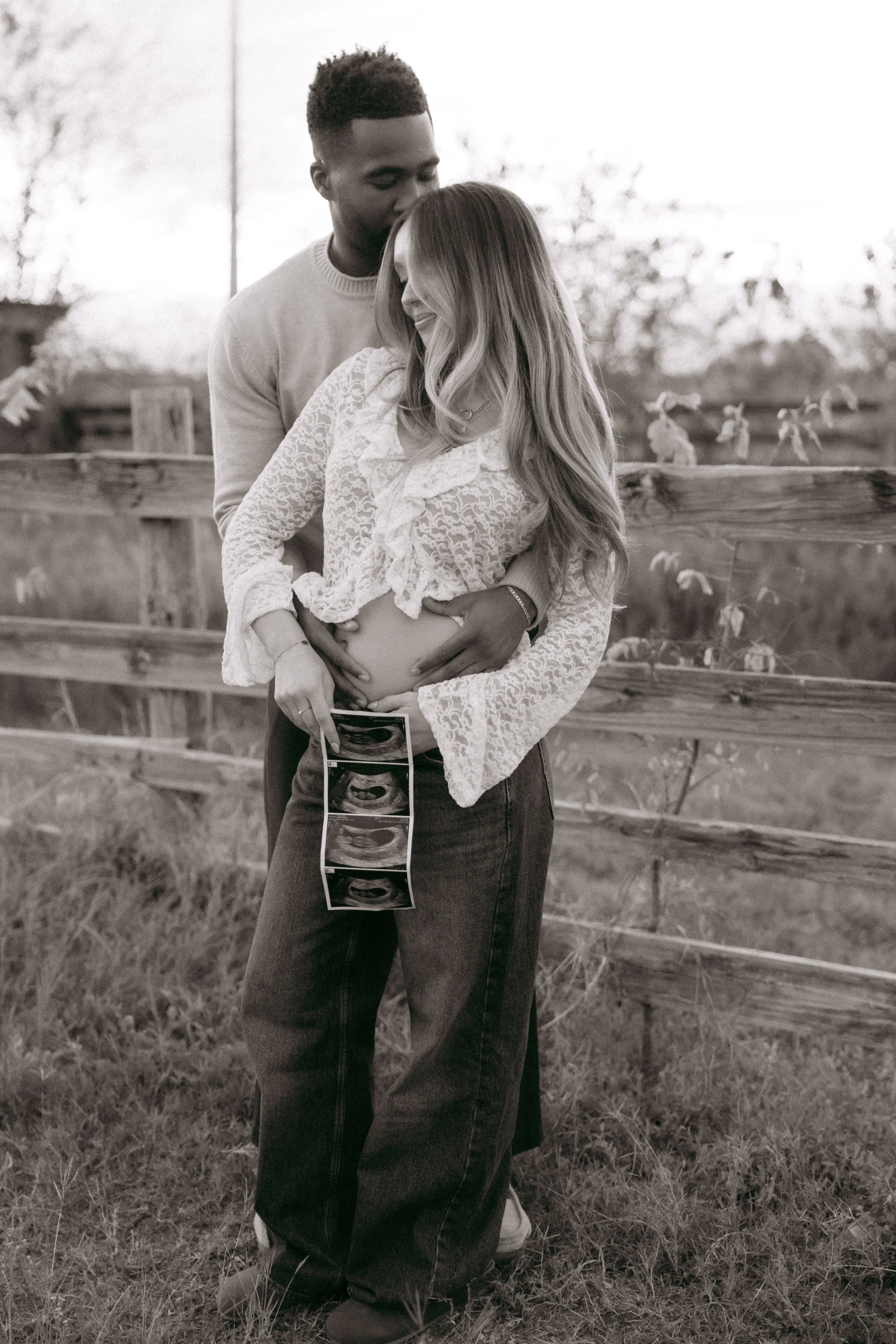 A pregnant woman holding ultrasound photos and a man standing behind her with his arms around her, outdoors with a wooden fence and trees in the background.