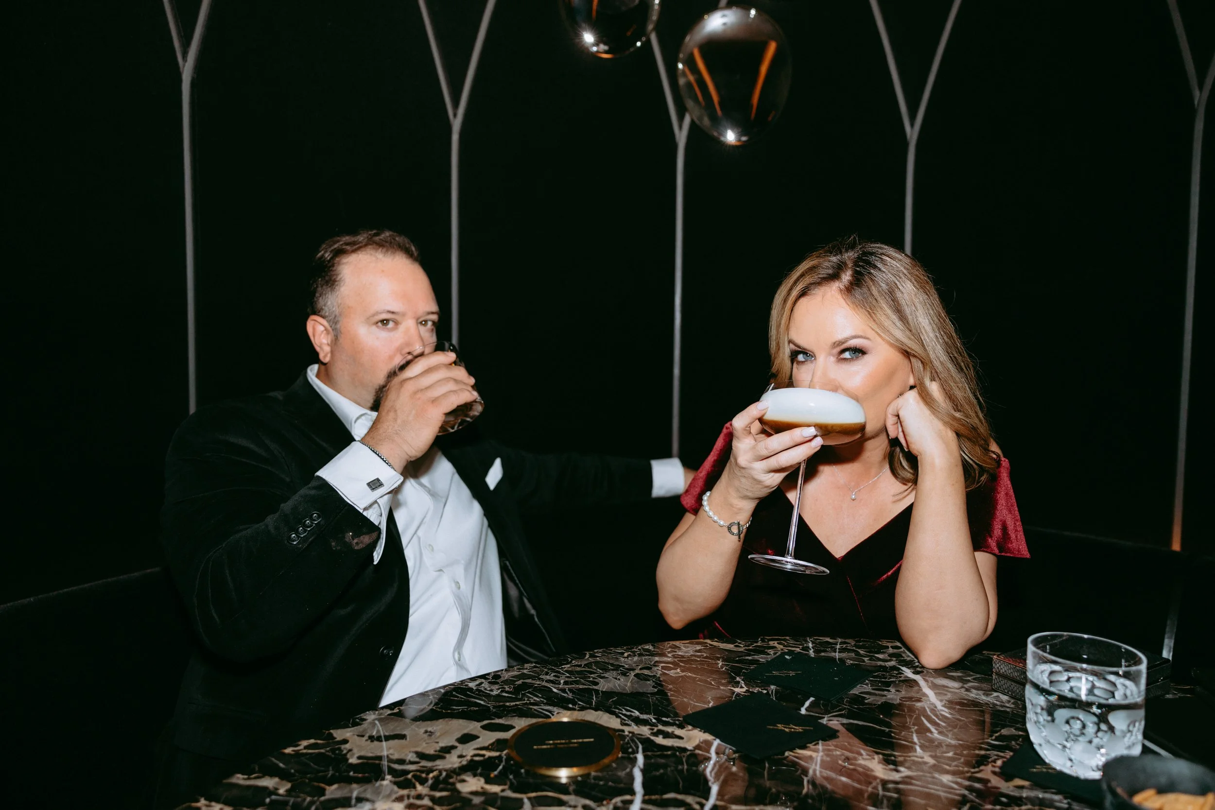 A man and woman sitting at a marble table in a dimly lit setting, each drinking from glasses, with the woman looking at the camera.
