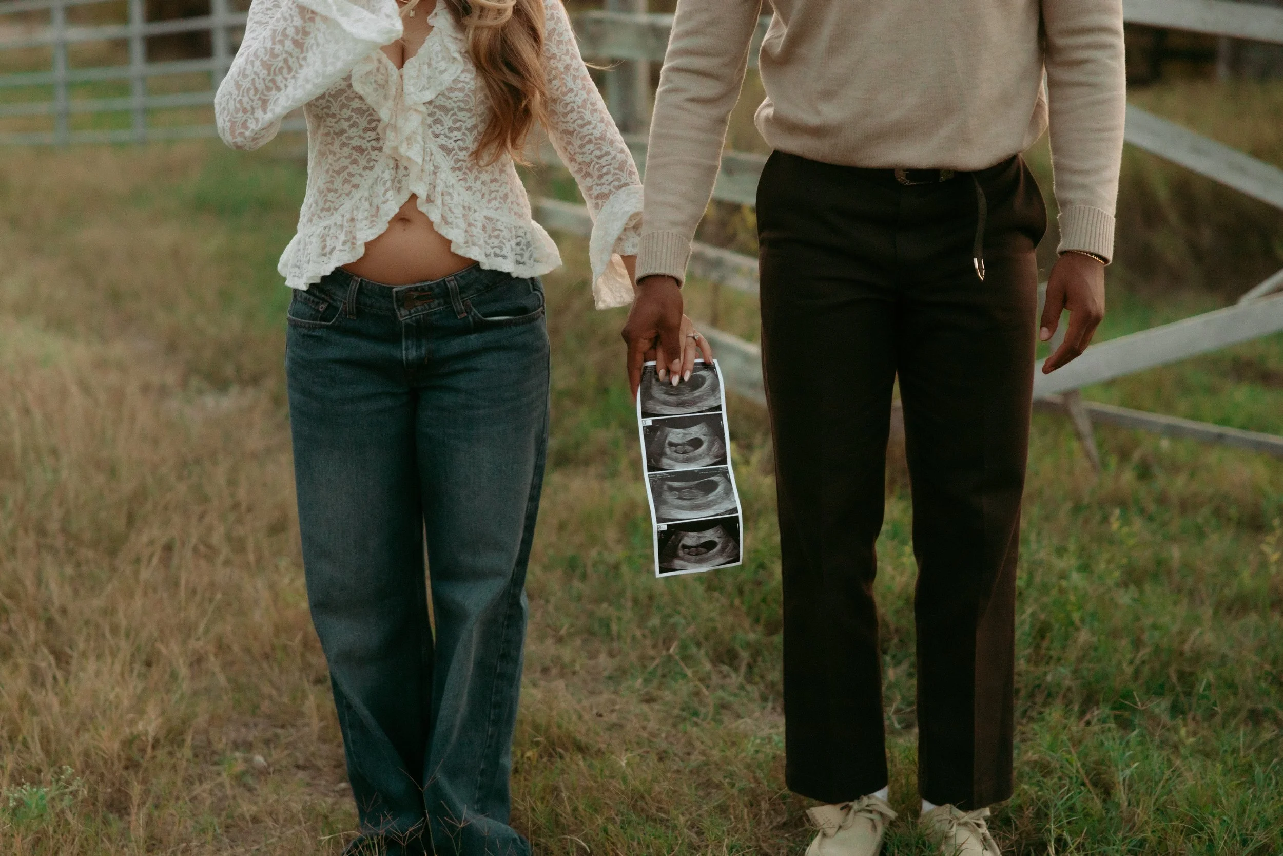 A couple holding ultrasound images outdoors on grass with a wooden fence in the background.