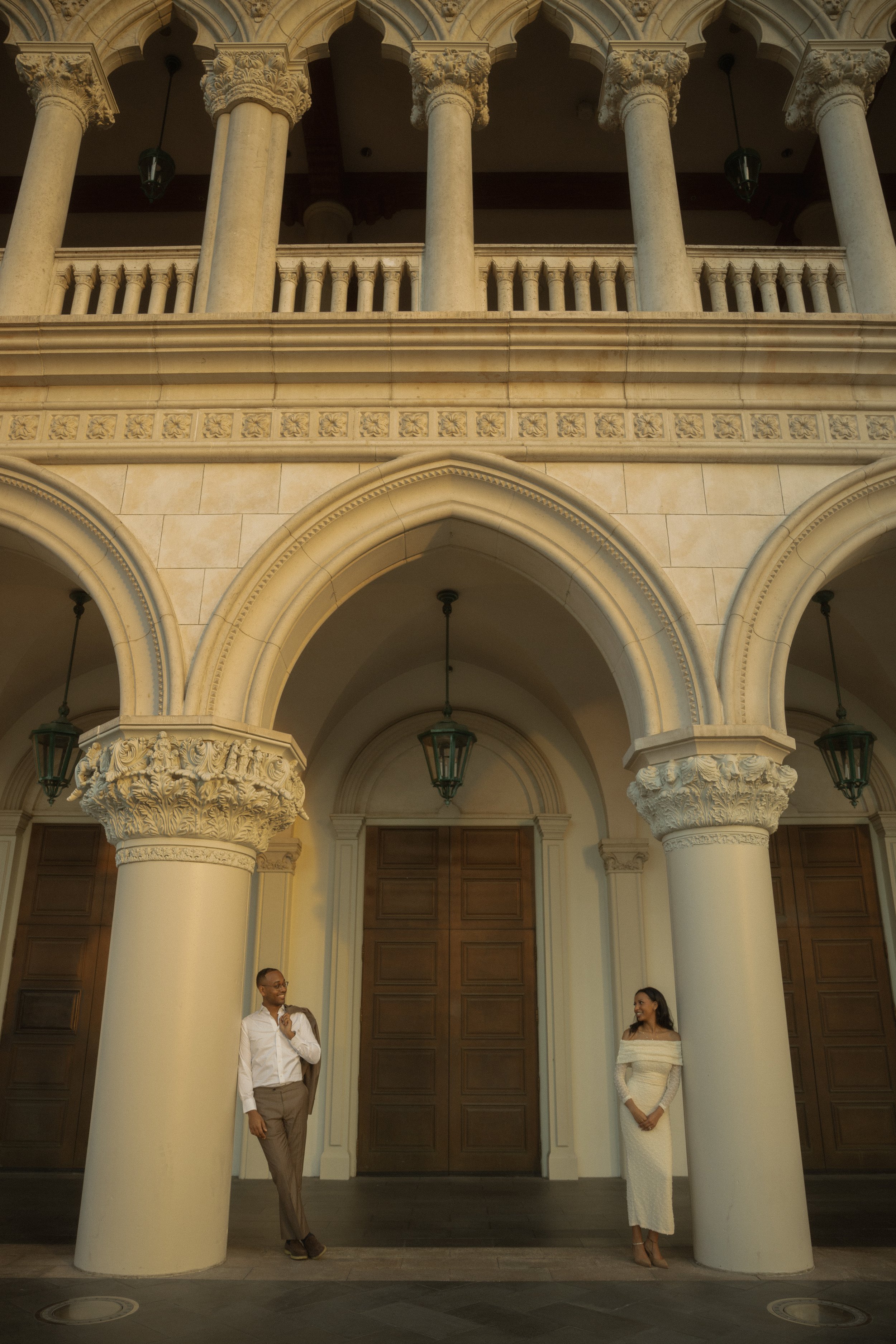 A man and woman standing apart in front of a grand, classical architectural building with large columns and arches. The man is on the left, wearing a white shirt and tan pants, holding a blazer over his shoulder. The woman is on the right, wearing a 