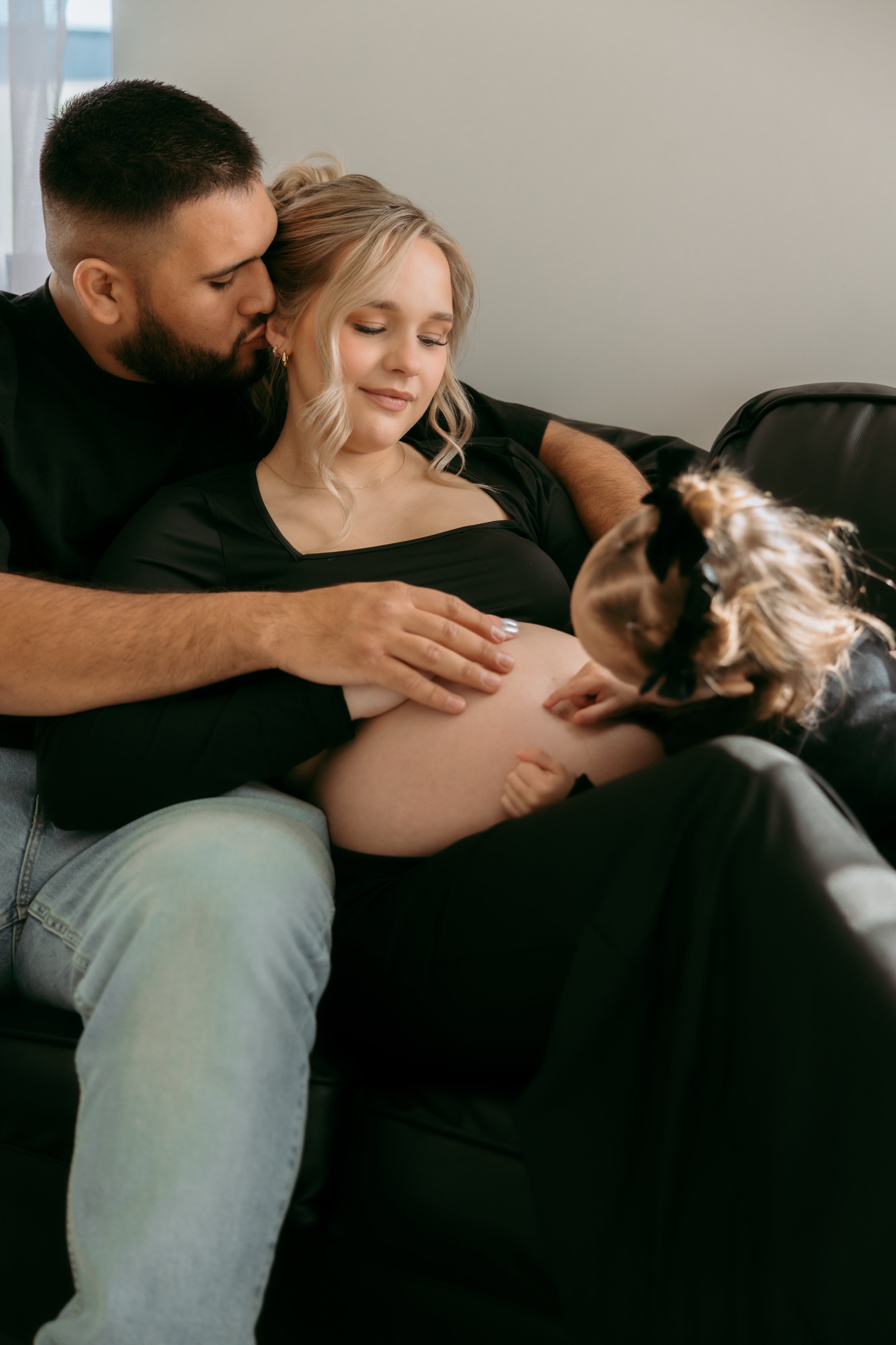 Pregnant woman with a man and a dog touching her belly, sitting on a couch in a living room.