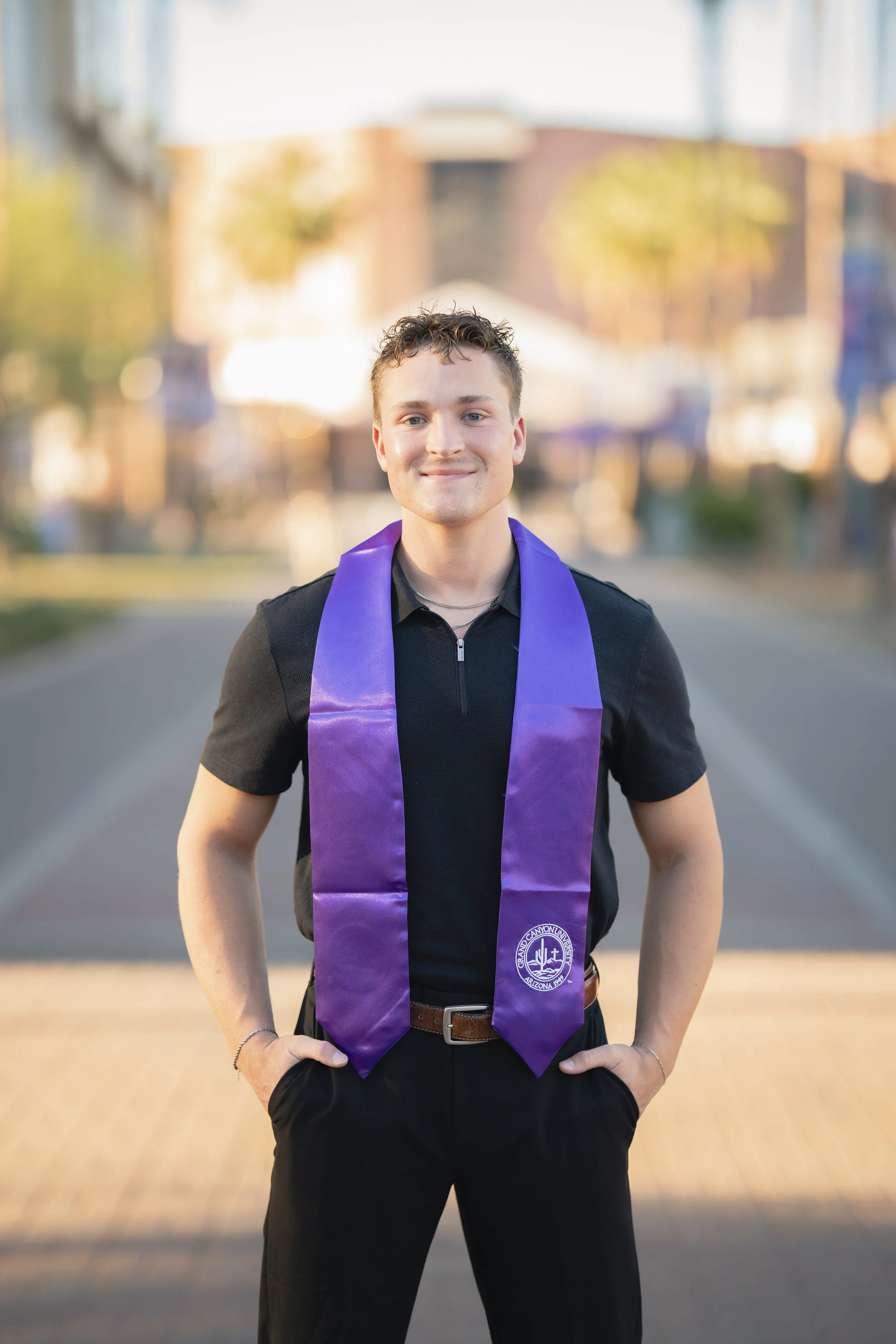 Young man in black shirt and pants wearing a purple graduation stole, standing outdoors with hands in pockets, smiling, with blurred trees and buildings in the background.