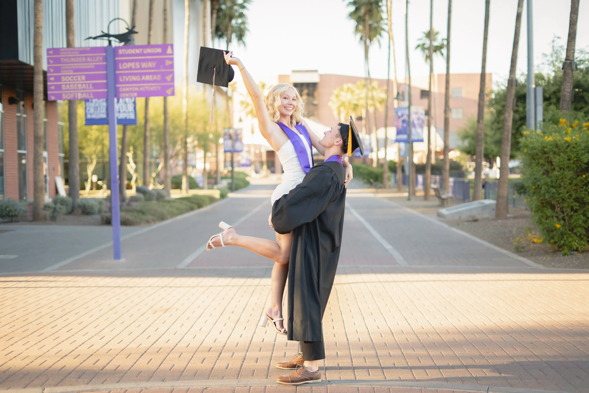 A young woman in a white dress and high heels is being lifted by a young man in a graduation cap and gown, celebrating graduation outdoors during daytime.