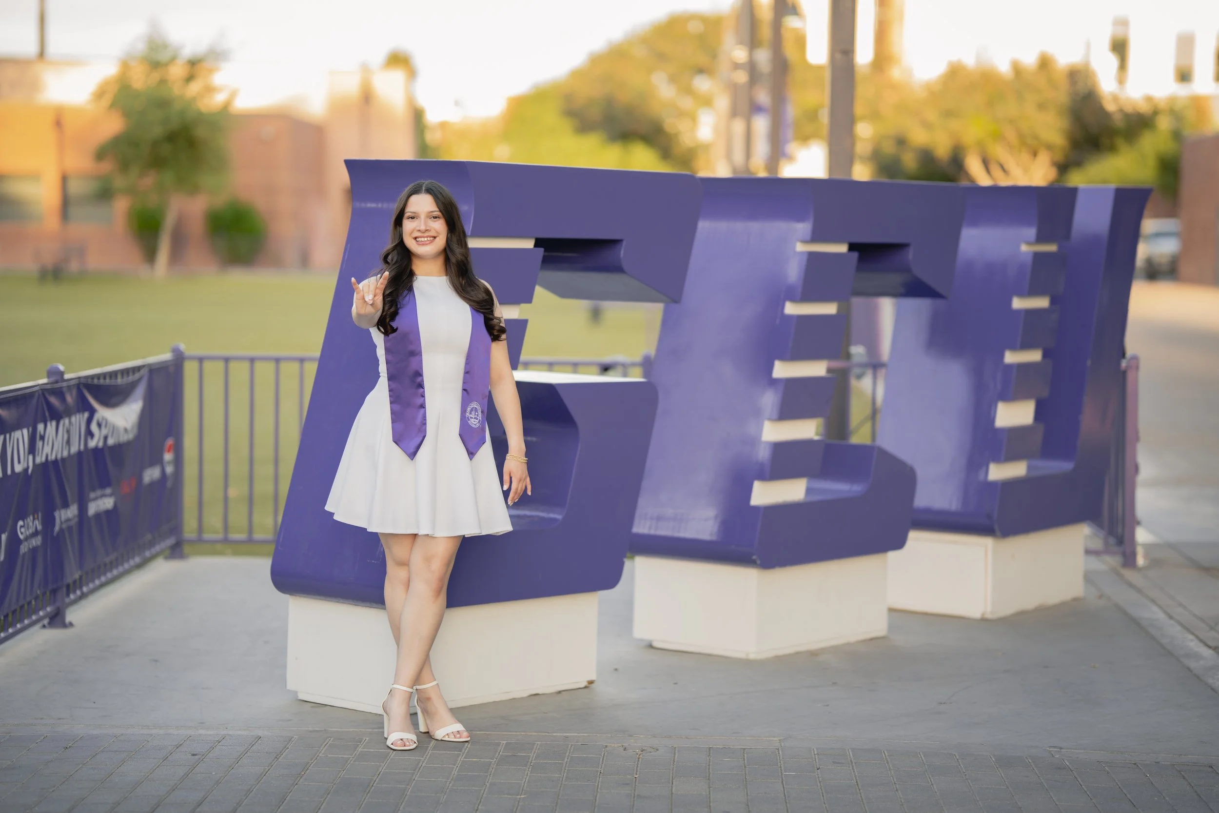 A young woman in a white dress and graduation stole standing next to a large blue 