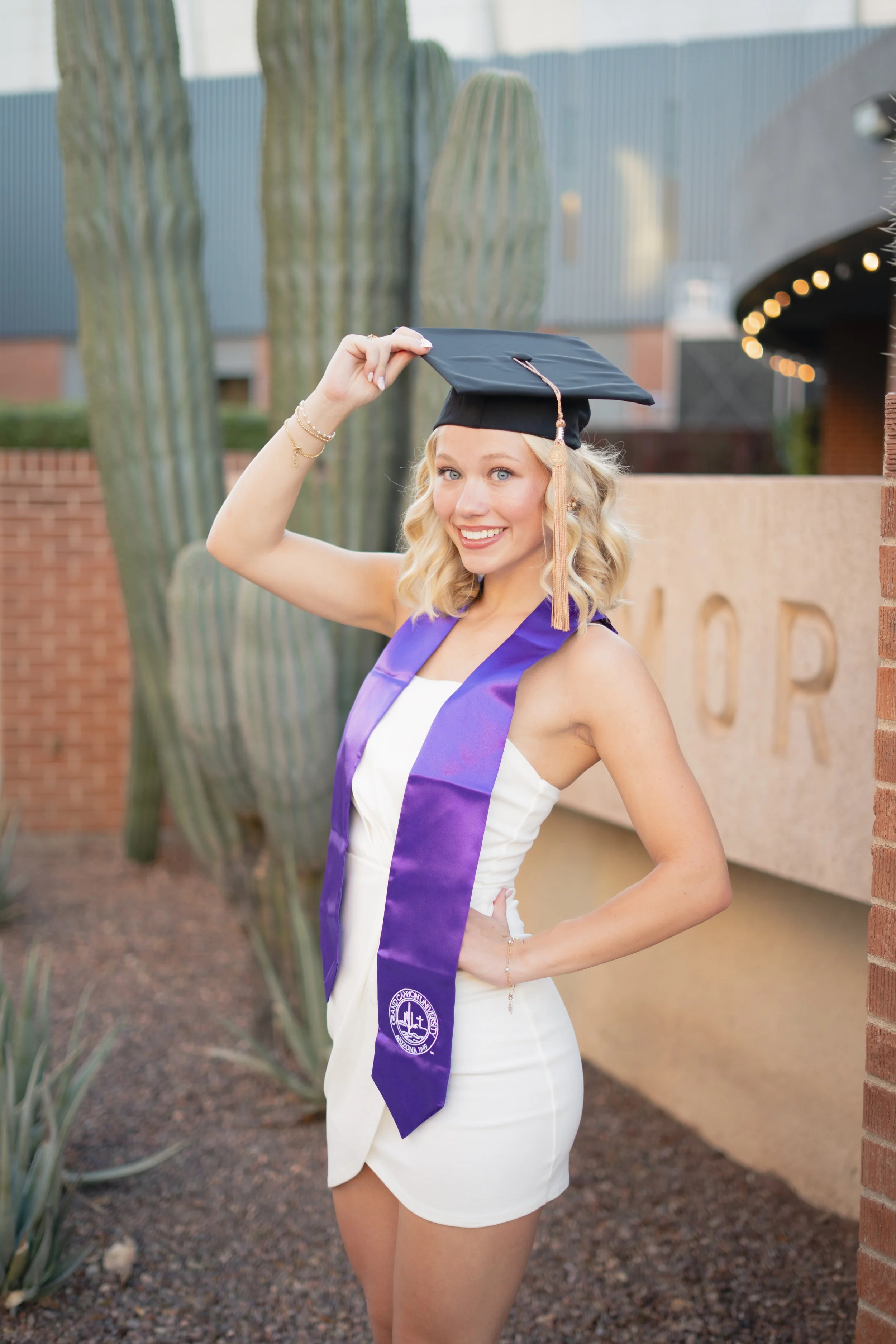 A young woman in a white dress wearing a purple sash and a graduation cap, smiling and holding the cap with her right hand, standing outdoors near cacti and a brick wall.