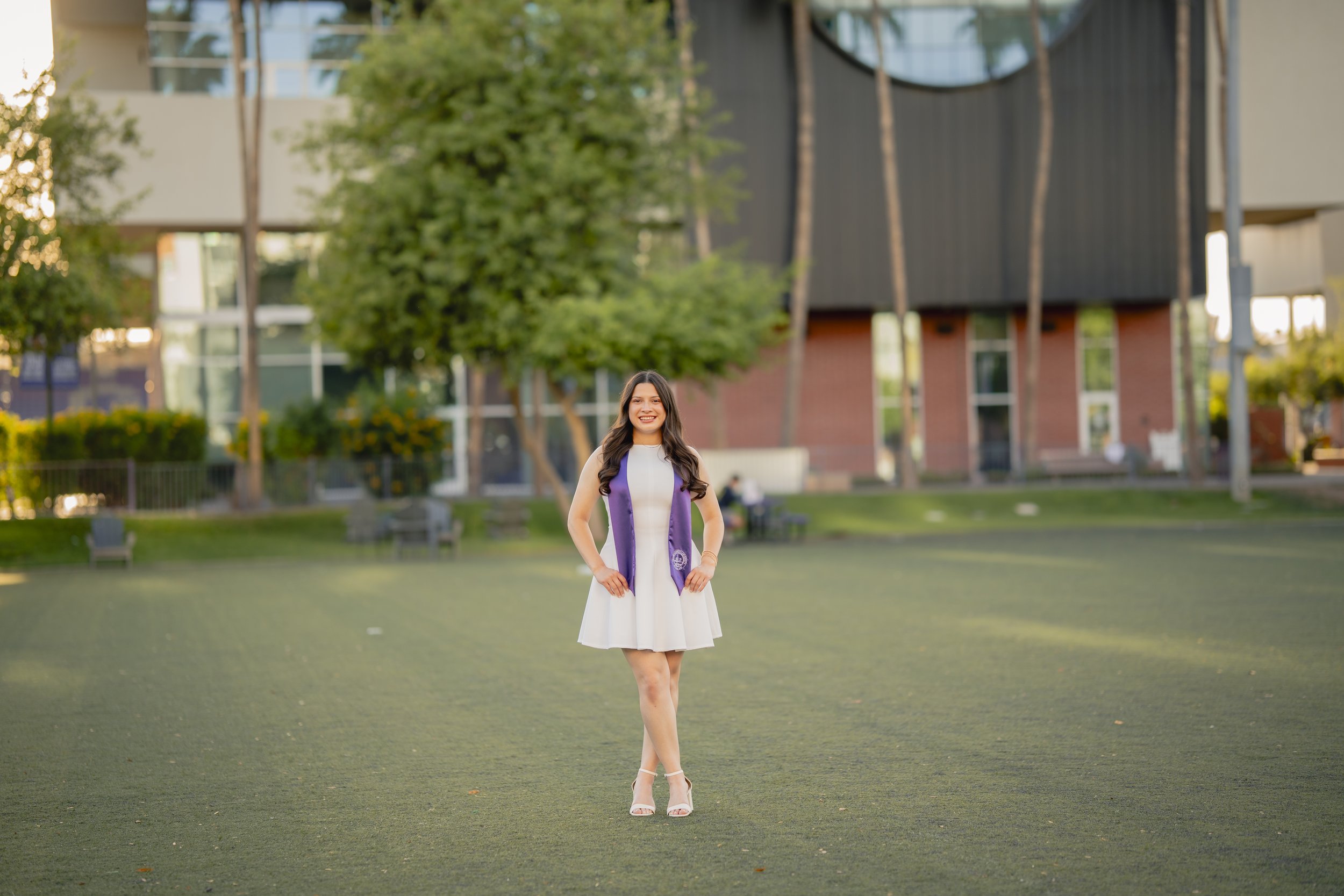 Young woman in a white dress and purple sash standing on a grassy field in a park with trees and modern buildings in the background.