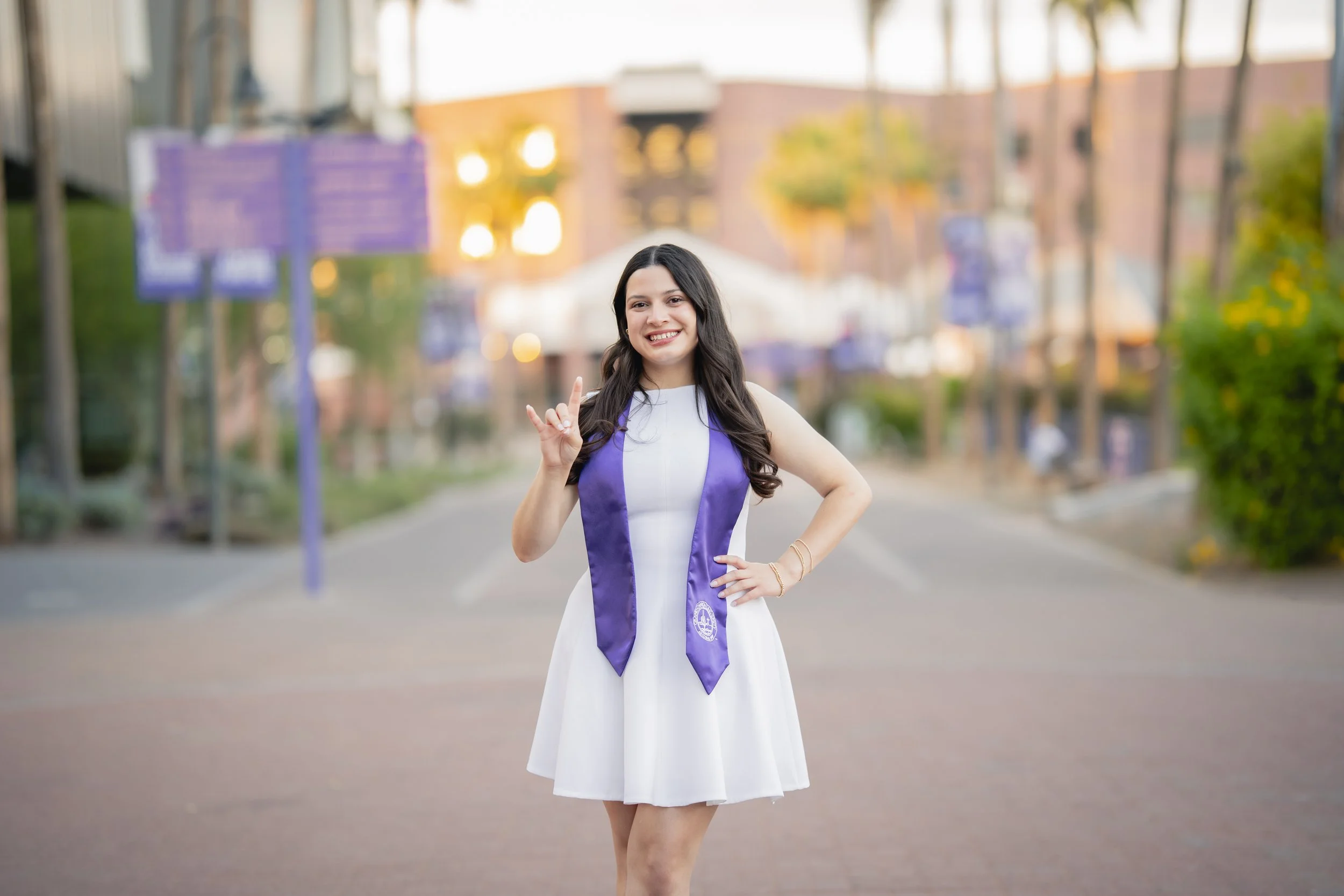 A young woman in a white dress and purple graduation stole smiling and making a hand gesture outdoors at sunset.