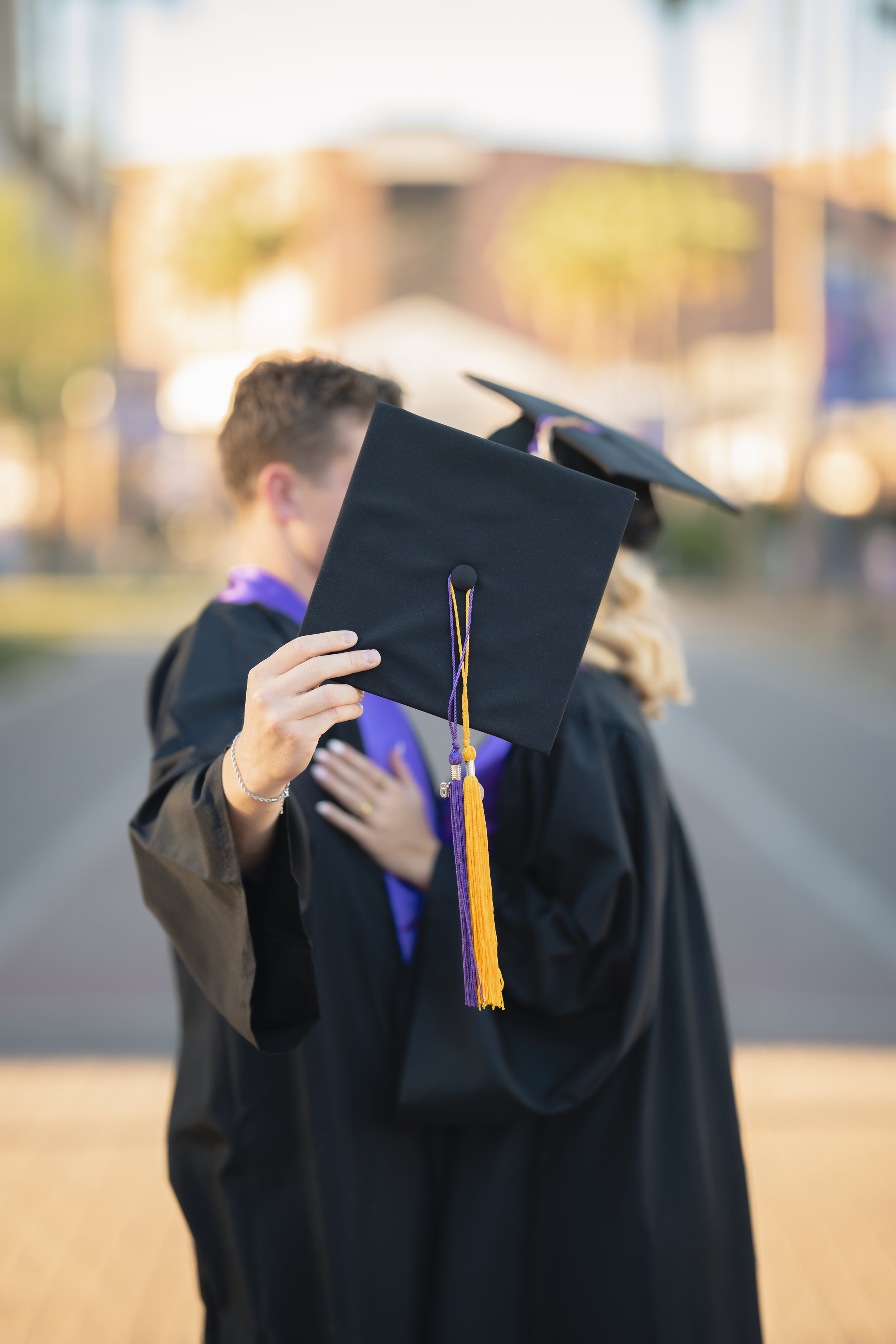 Graduate holding up graduation cap during ceremony, with other graduate in background, outdoor setting, autumn trees and buildings in distance.