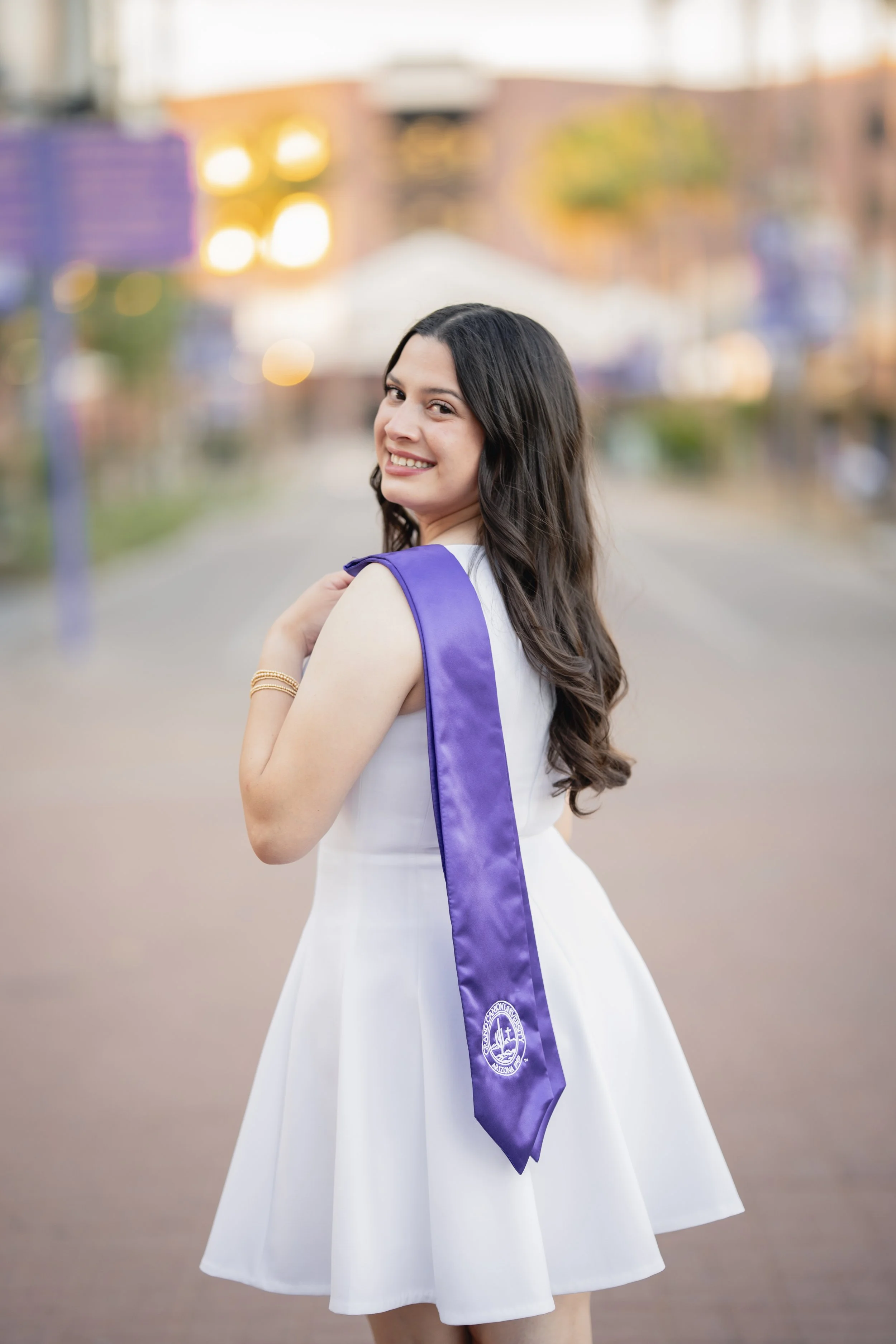 A young woman in a white dress with a purple sash over her shoulder, smiling at the camera outdoors during sunset.