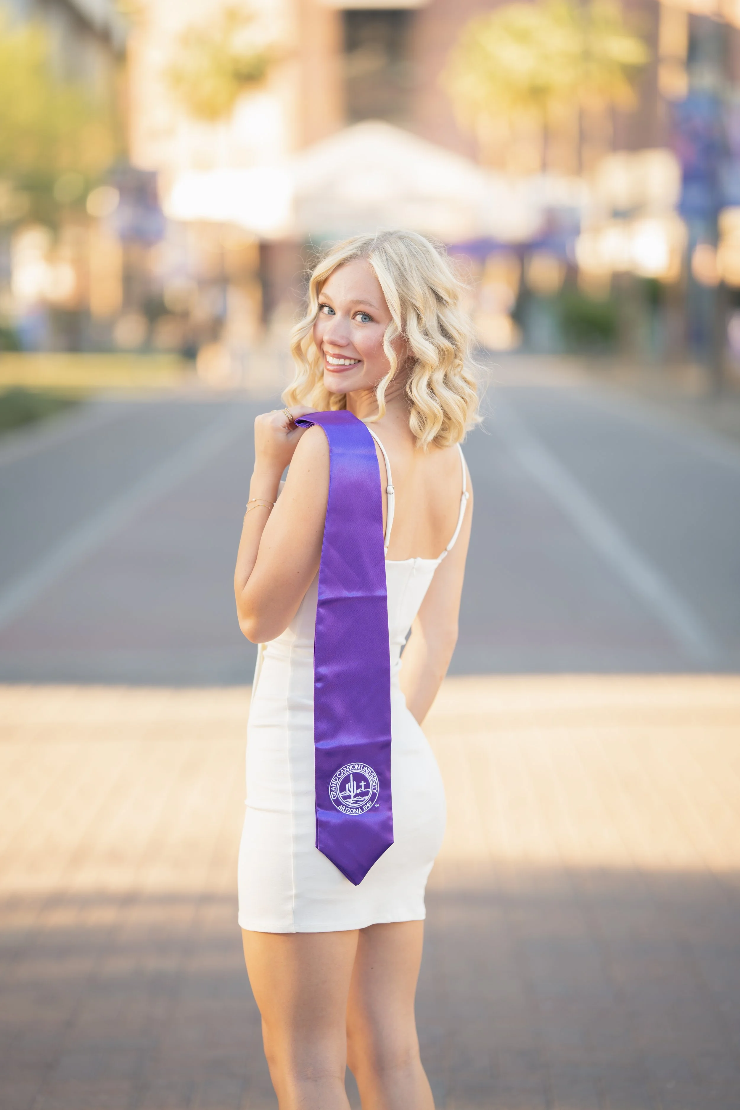 A young woman with blonde curly hair wearing a white dress, smiling and looking over her shoulder, with a purple sash hanging off her shoulder, outdoors during sunset.