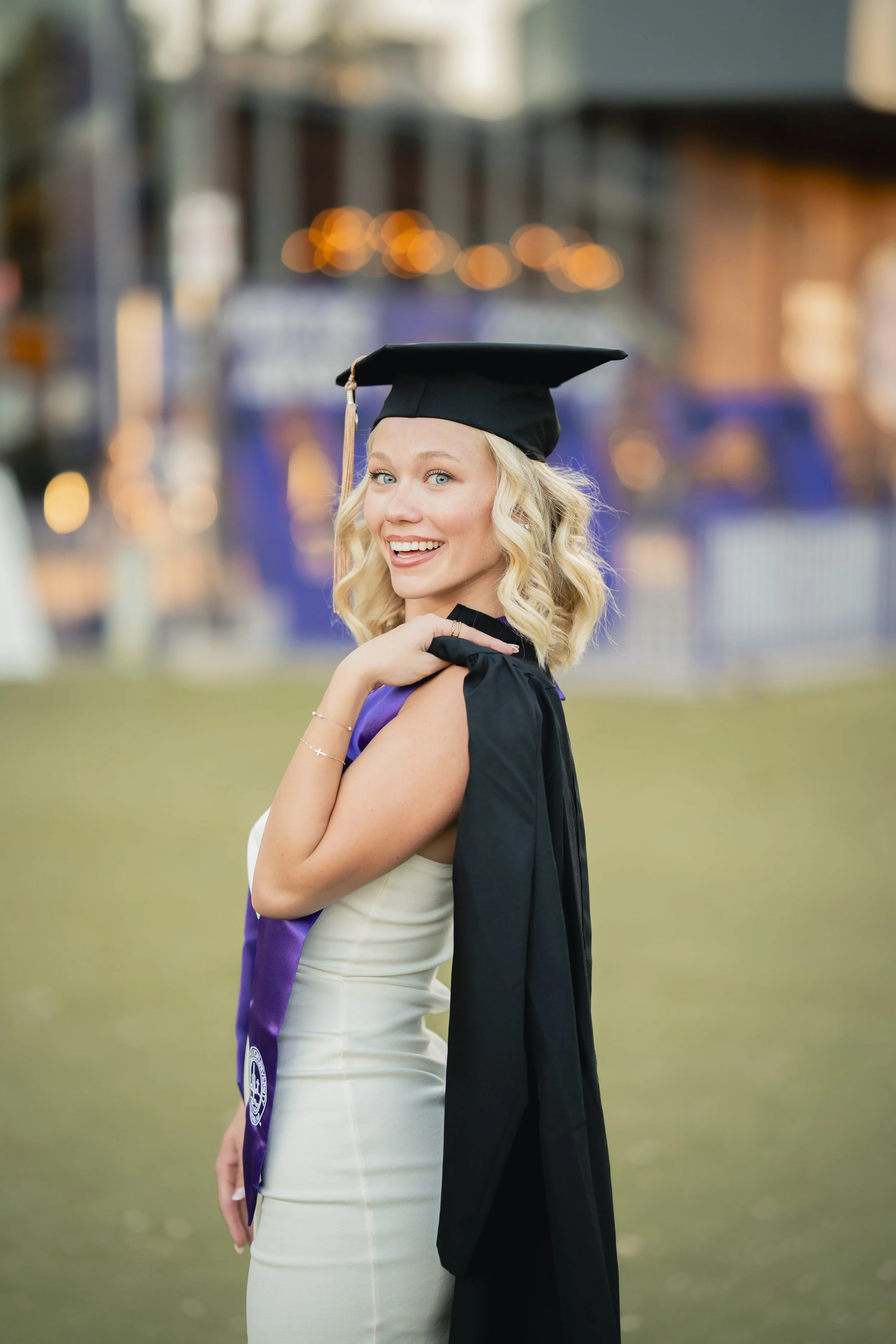 A young woman in a graduation cap and gown, smiling and posing outdoors on a grassy area, with colorful lights and buildings blurred in the background.