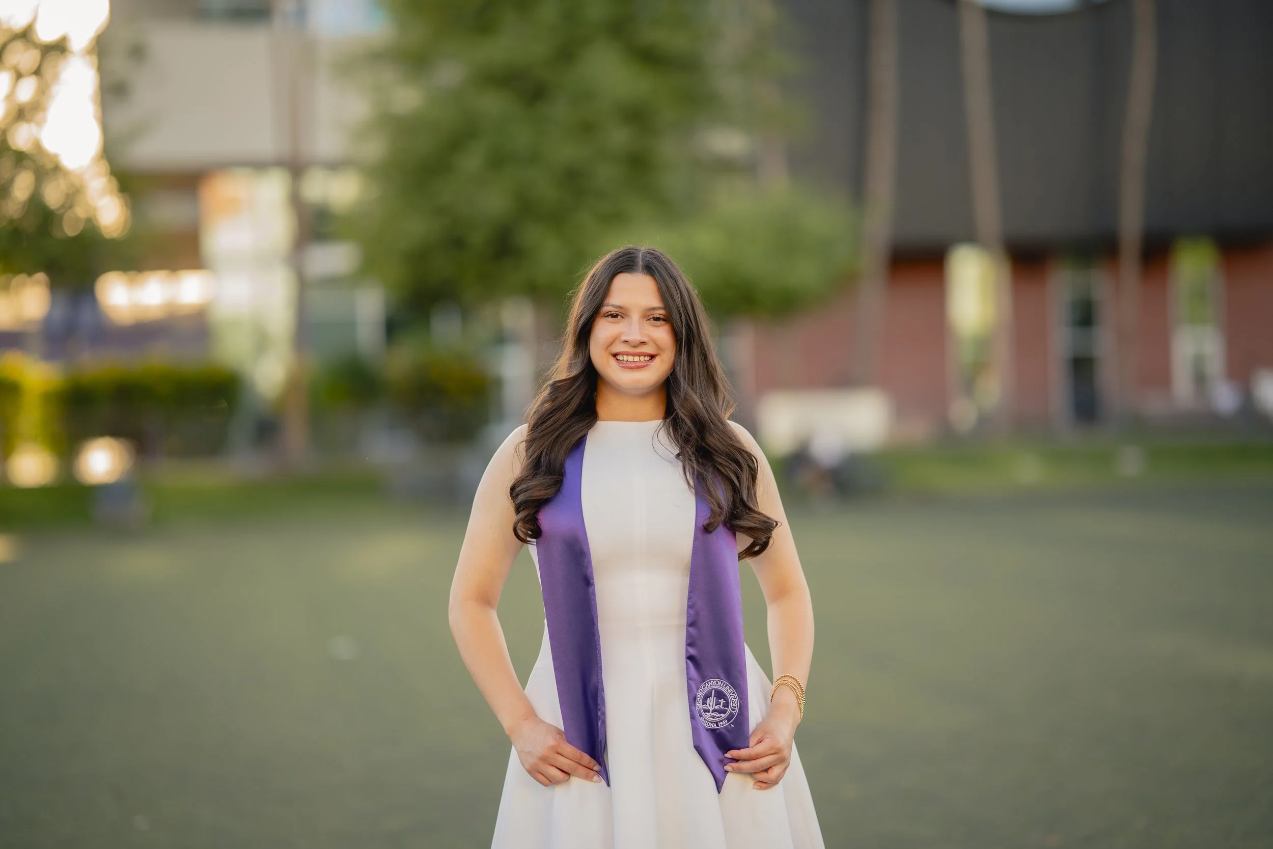 Young woman in a white dress with purple sash, smiling outdoors in a park setting.