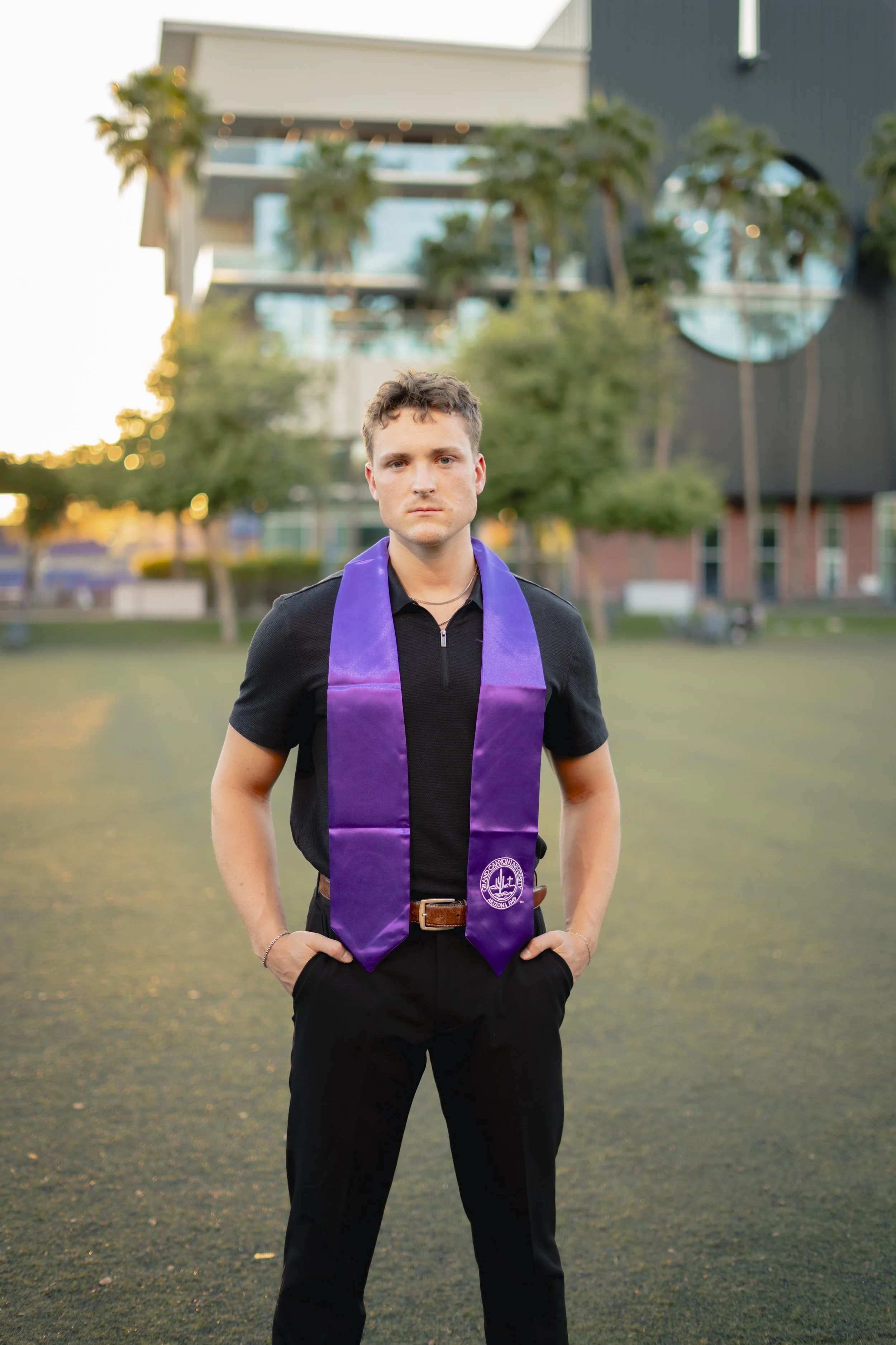 Young man in black shirt and pants with a purple stole standing outdoors on a grassy area during sunset.