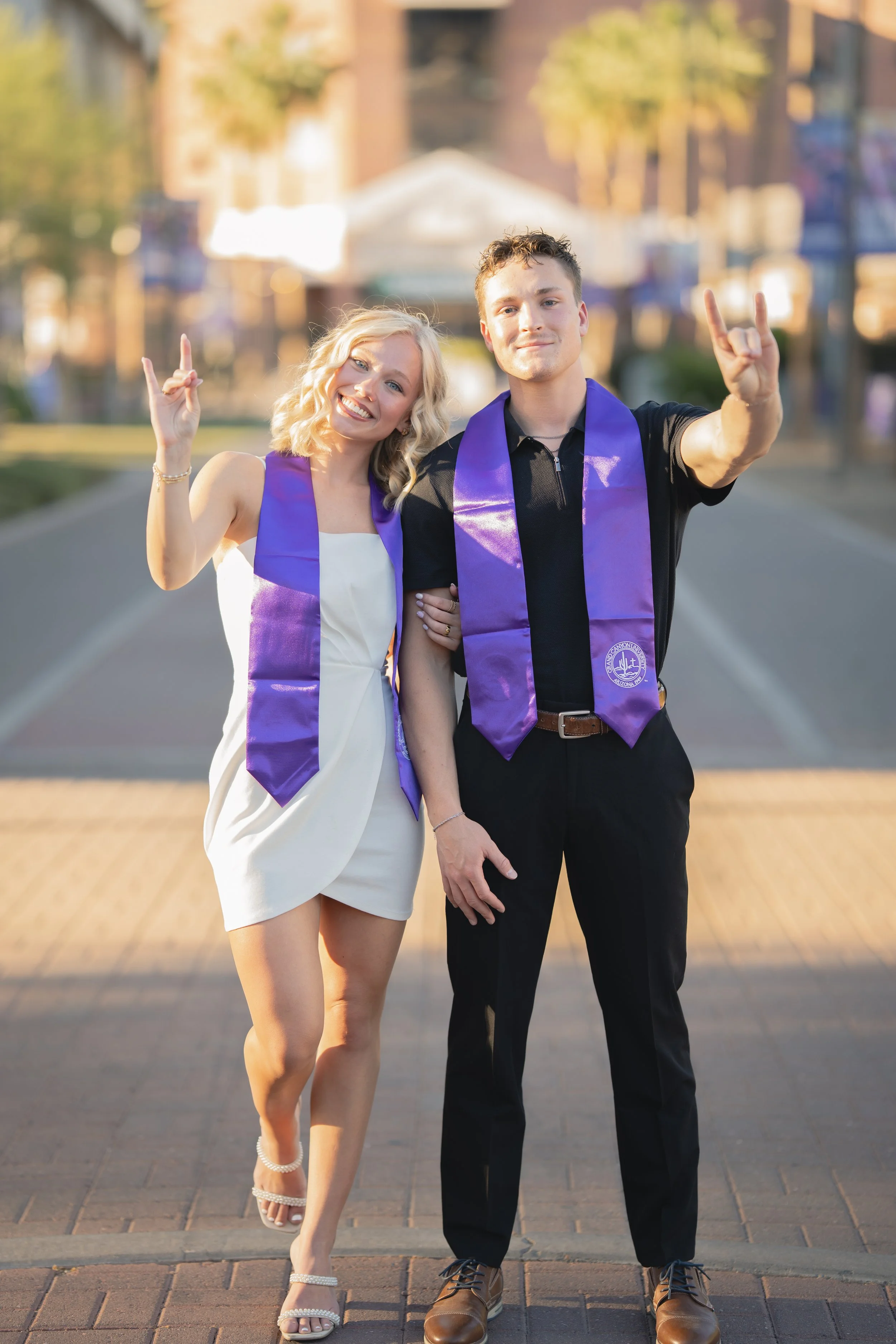 A young couple at graduation celebration wearing purple sashes, smiling, and making celebratory gestures in an outdoor urban setting during sunset.