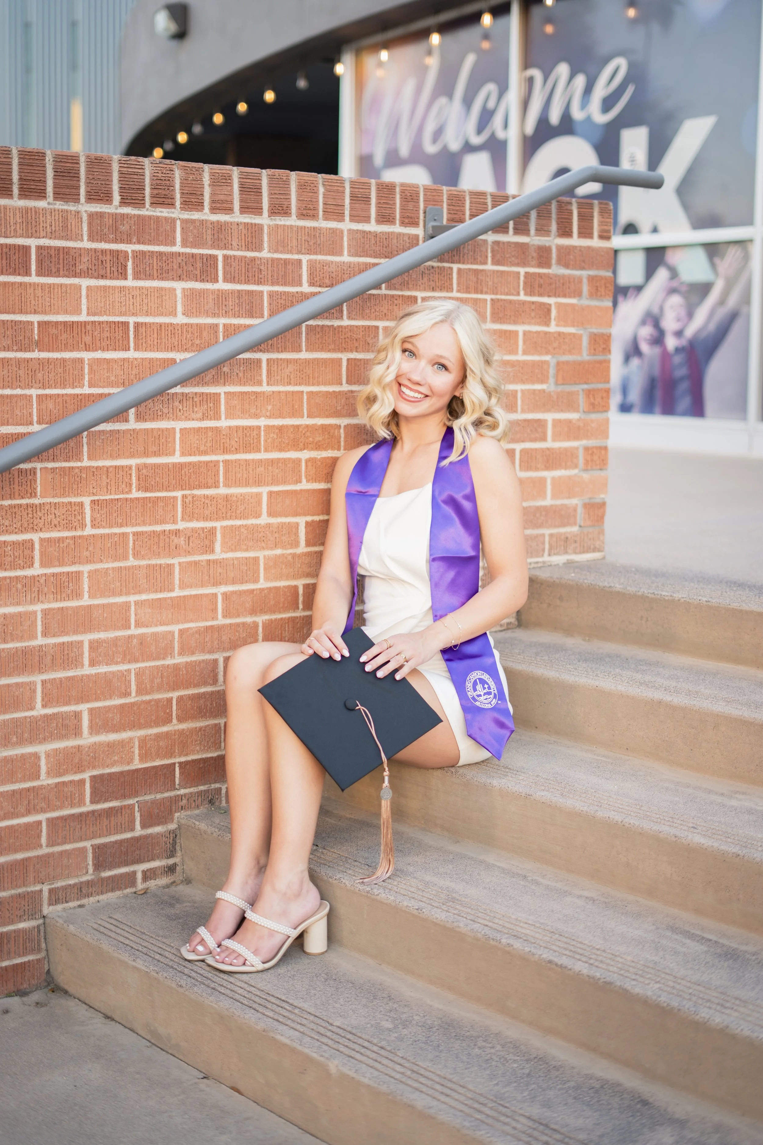 A young woman with blonde, curly hair is sitting on outdoor steps, smiling while holding a graduation cap, wearing a white dress and a purple graduation stole.
