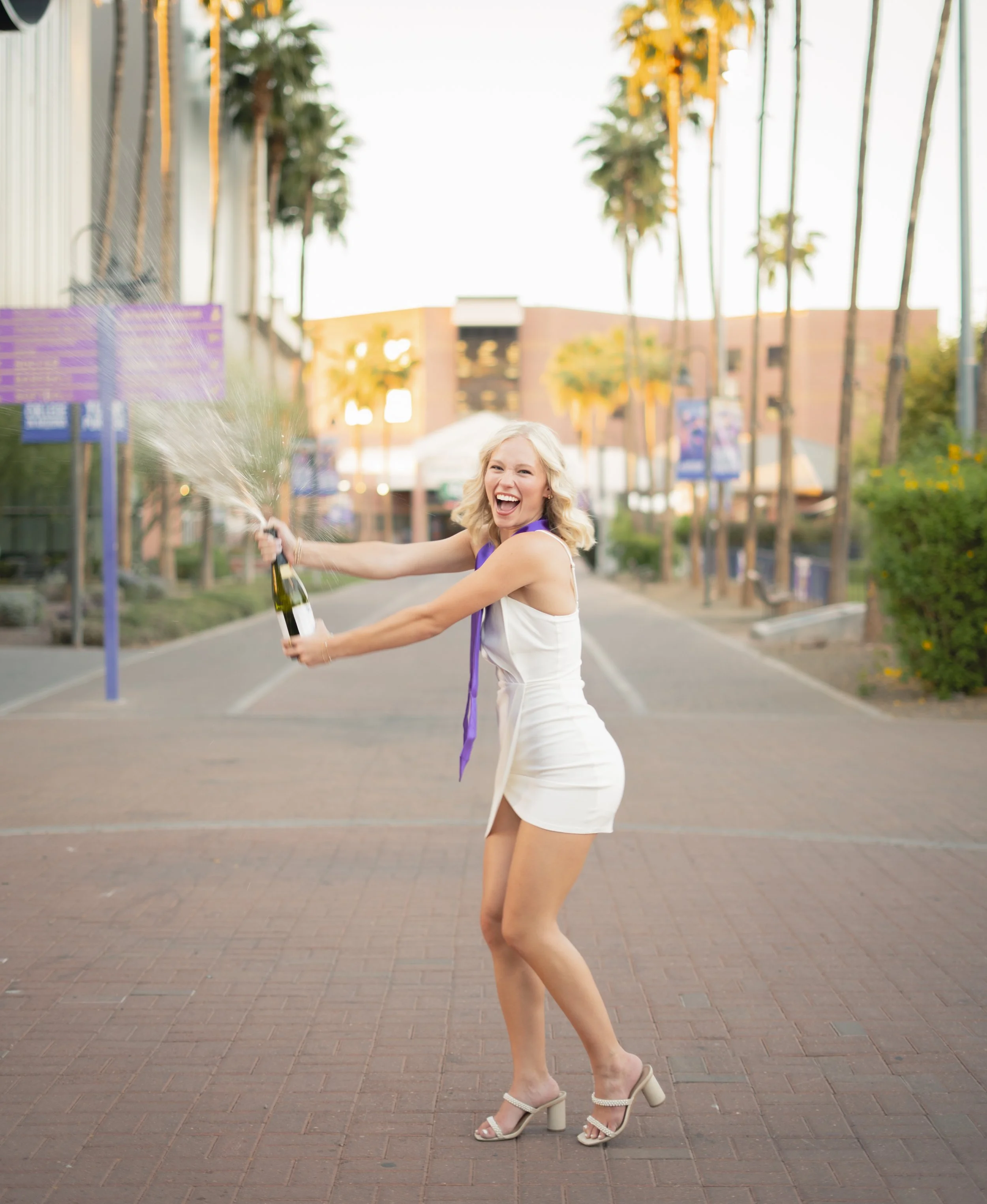 A woman in a white dress and high heels celebrating outdoors while opening a champagne bottle, with champagne spraying out, on a brick pathway lined with palm trees.