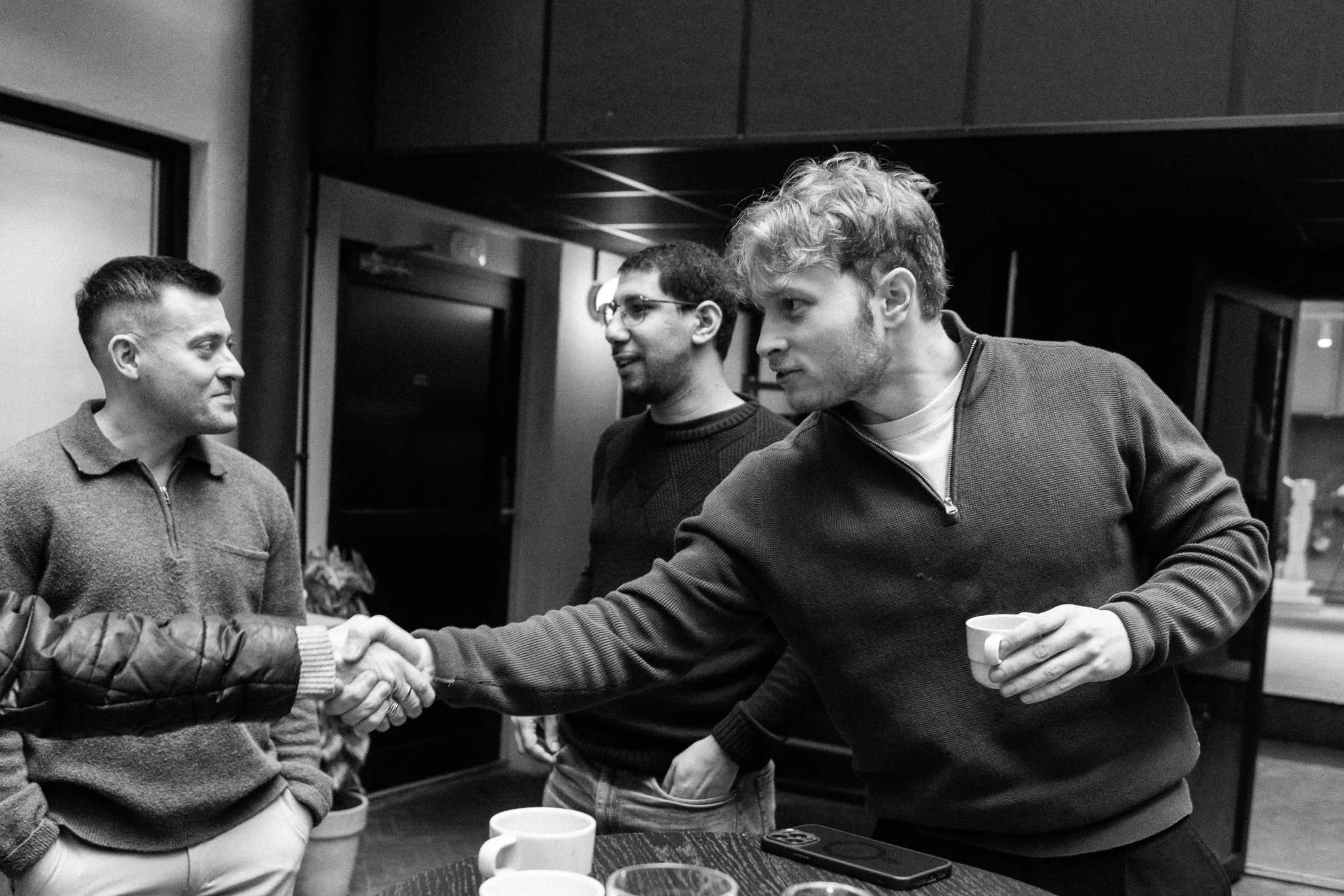 Four men in a room, two of them shaking hands, with cups on the table in front of them.