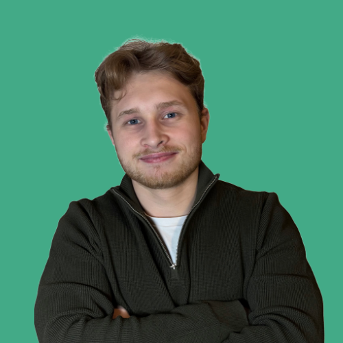 A young man with light brown hair and a beard, smiling, wearing a black jacket and white shirt, standing with arms crossed against a green background.