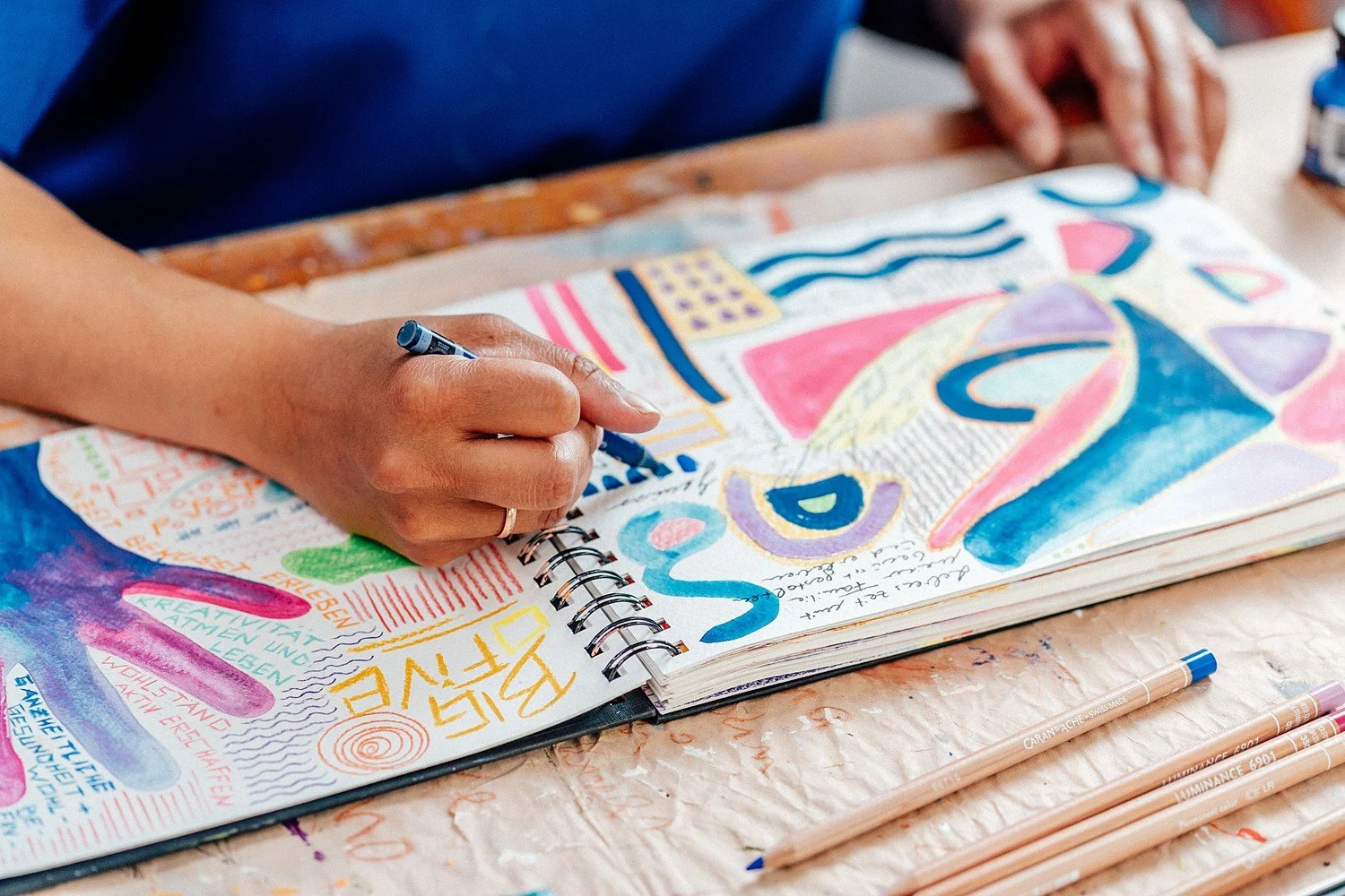 Person zeichnet bunte abstrakte Kunstwerke in einem Skizzenbuch, umgeben von Markern auf einem Tisch.