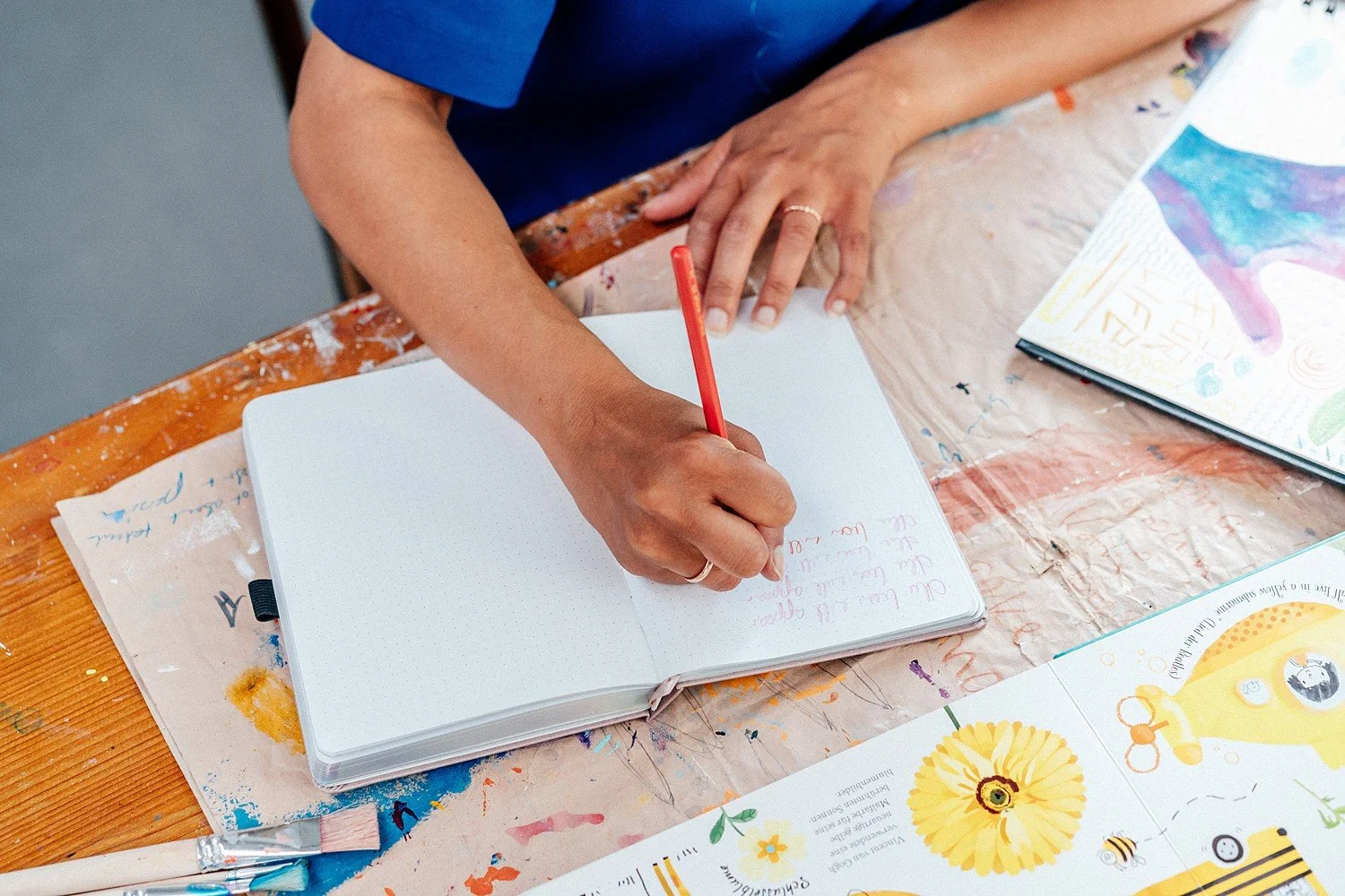 Eine Person schreibt mit einem roten Stift in einem weißen, gepunkteten Notizbuch. Der Tisch ist mit bunten Farben und Acrylfarben bedeckt, und es liegen Malbücher sowie eine Pinsel auf dem Tisch.