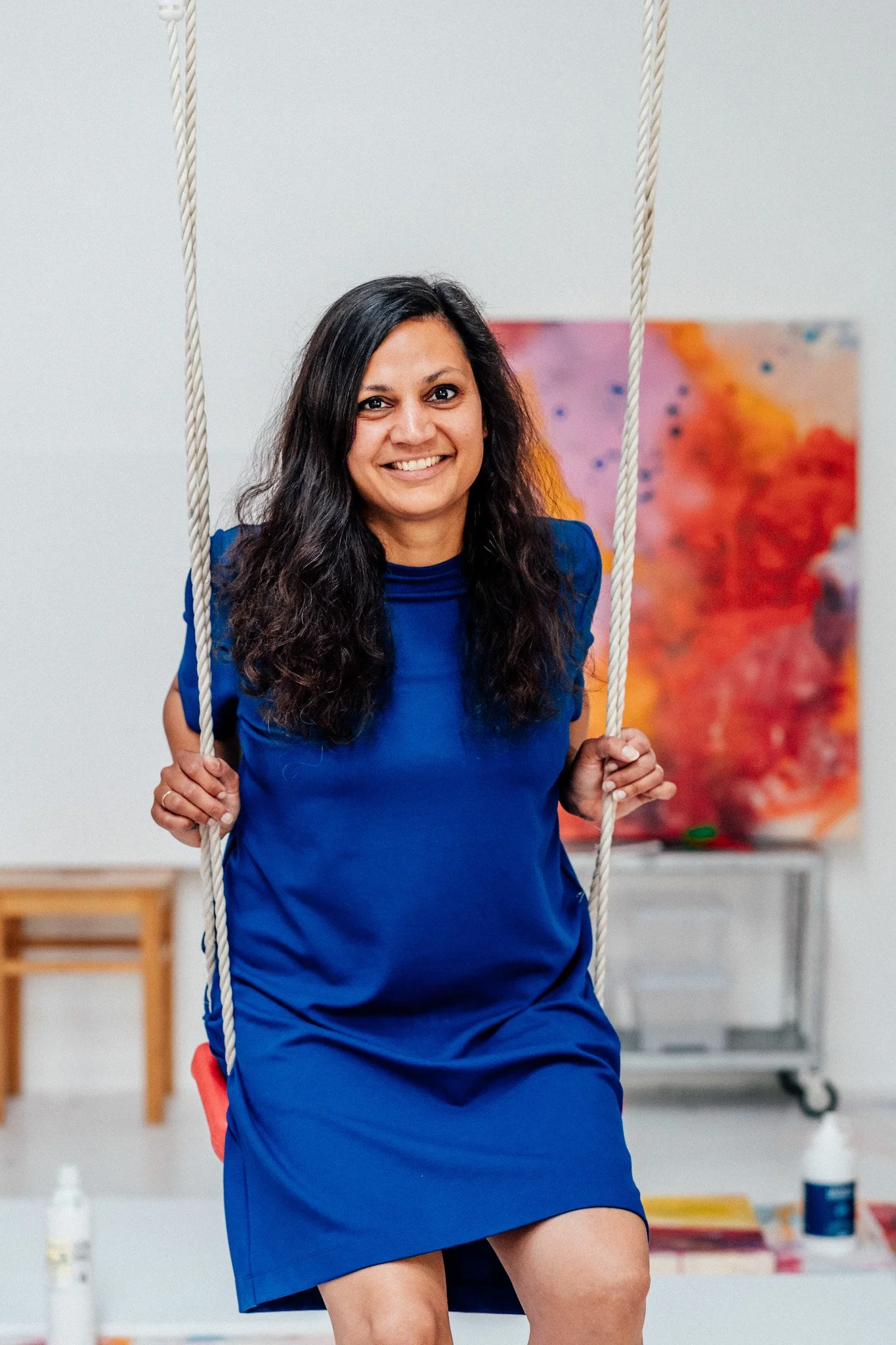 Eine lächelnde Frau mit dunklen, lockigen Haaren in einem blauen Kleid sitzt auf einer Schaukel im Kunststudio.