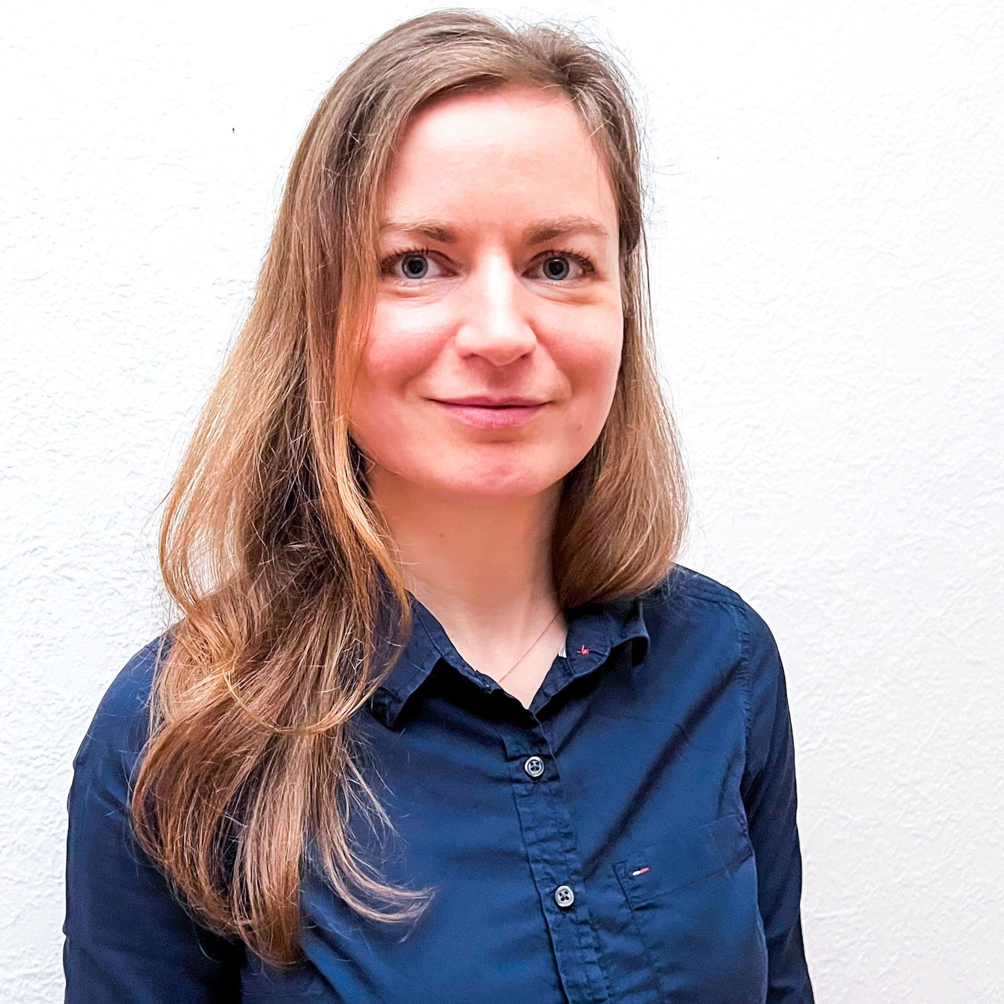 Nahaufnahme eines lachenden Mädchens mit langen, hellbraunen Haaren, blauen Augen und einem blauen Hemd vor einem weißen Hintergrund.
