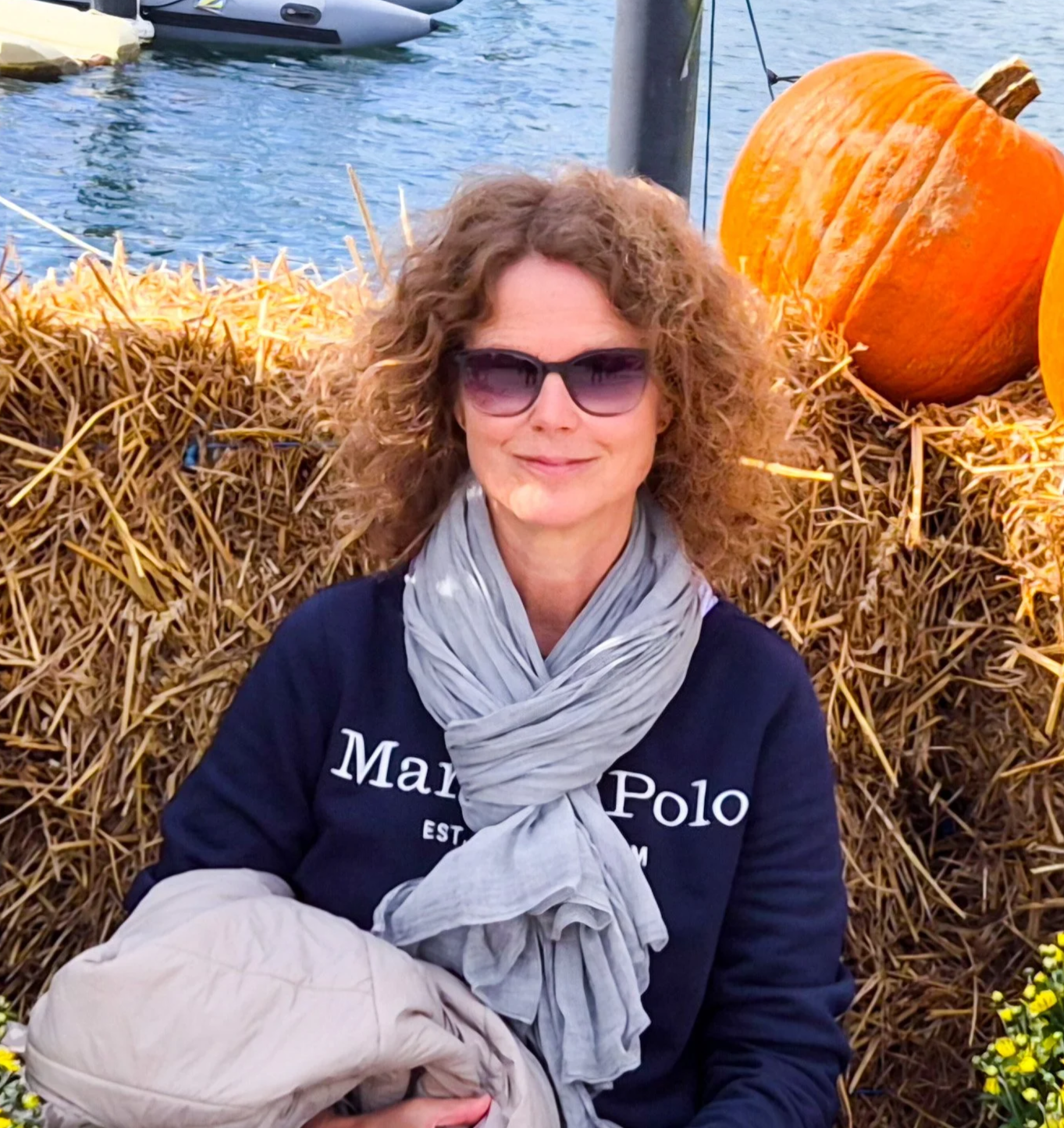 Eine Frau mit lockigem Haar, Sonnenbrille und einem dunkelblauen Pullover mit weißer Schrift, die auf einer Strohmauer sitzt, im Hintergrund Kürbisse und Wasser.