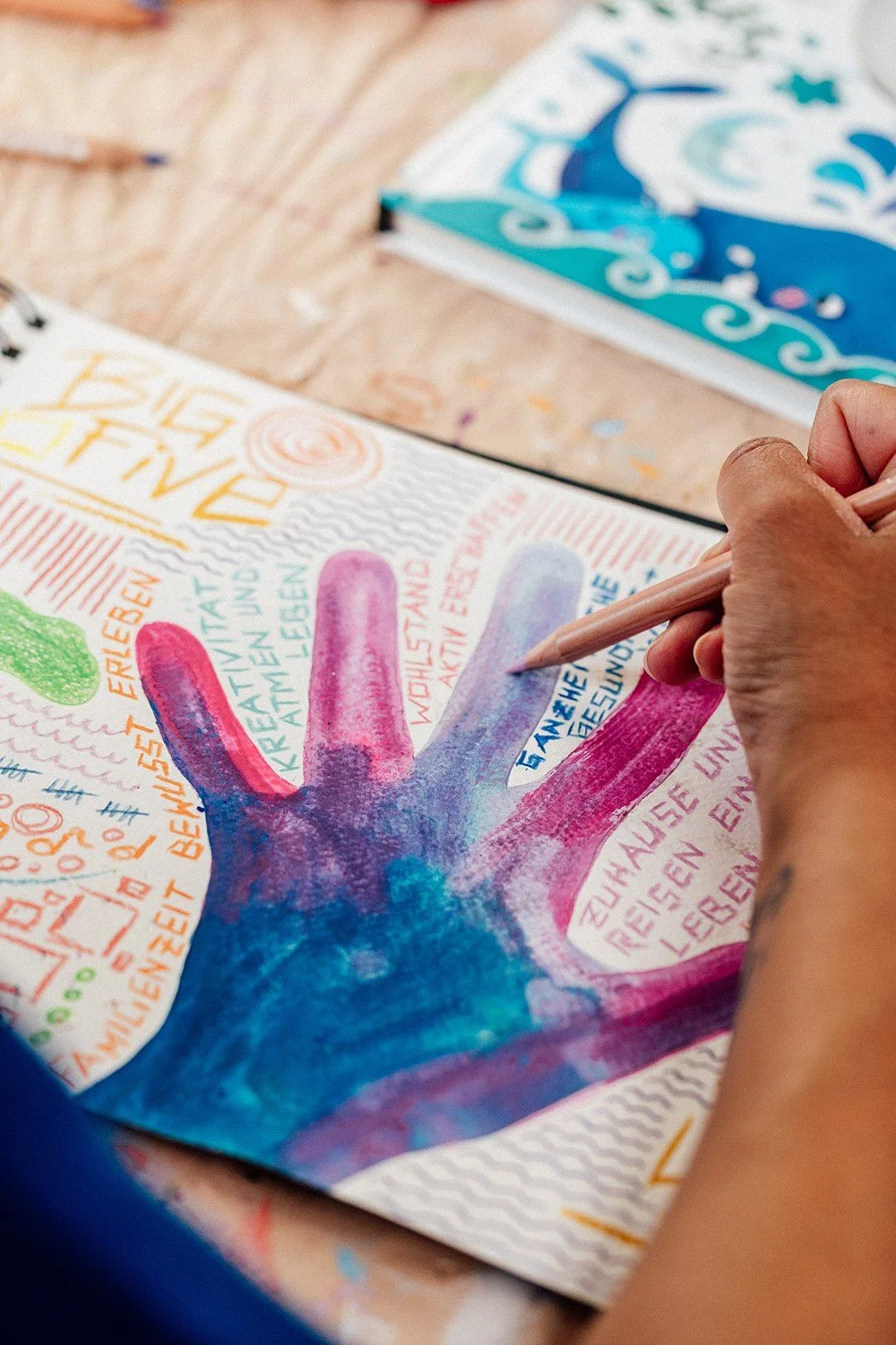 Hand, die eine bunte Handabdruckzeichnung mit Wasserfarben ausmalt, auf einem Tisch mit anderen Kunstmaterialien.