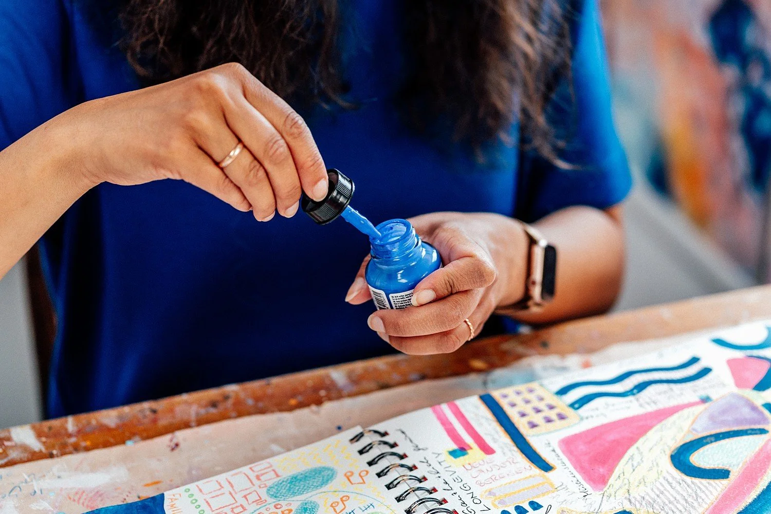 Eine Frau, die blauen Nagellack aufträgt, hält eine Flasche mit blauer Farbe und einen Tropfer. Sie sitzt an einem Tisch, auf dem ein bunt bemalter Zeichenblock liegt.