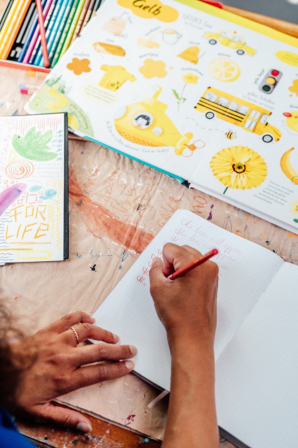 Eine Person schreibt in ein Notizbuch mit rotem Stift auf einem Tisch, umgeben von bunten Buntstiften, einem Malbuch mit Gelb-Thema, und einer Zeichnung mit dem Wort 'FOR LIFE'.
