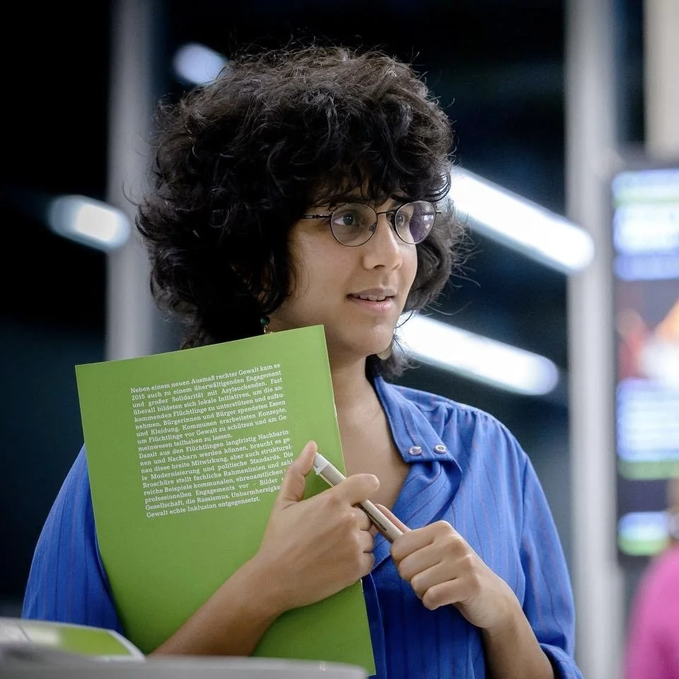 Junge Frau mit Brille und lockigem Haar hält eine grüne Broschüre und einen Stift in der Hand, steht in einer öffentlichen Umgebung.