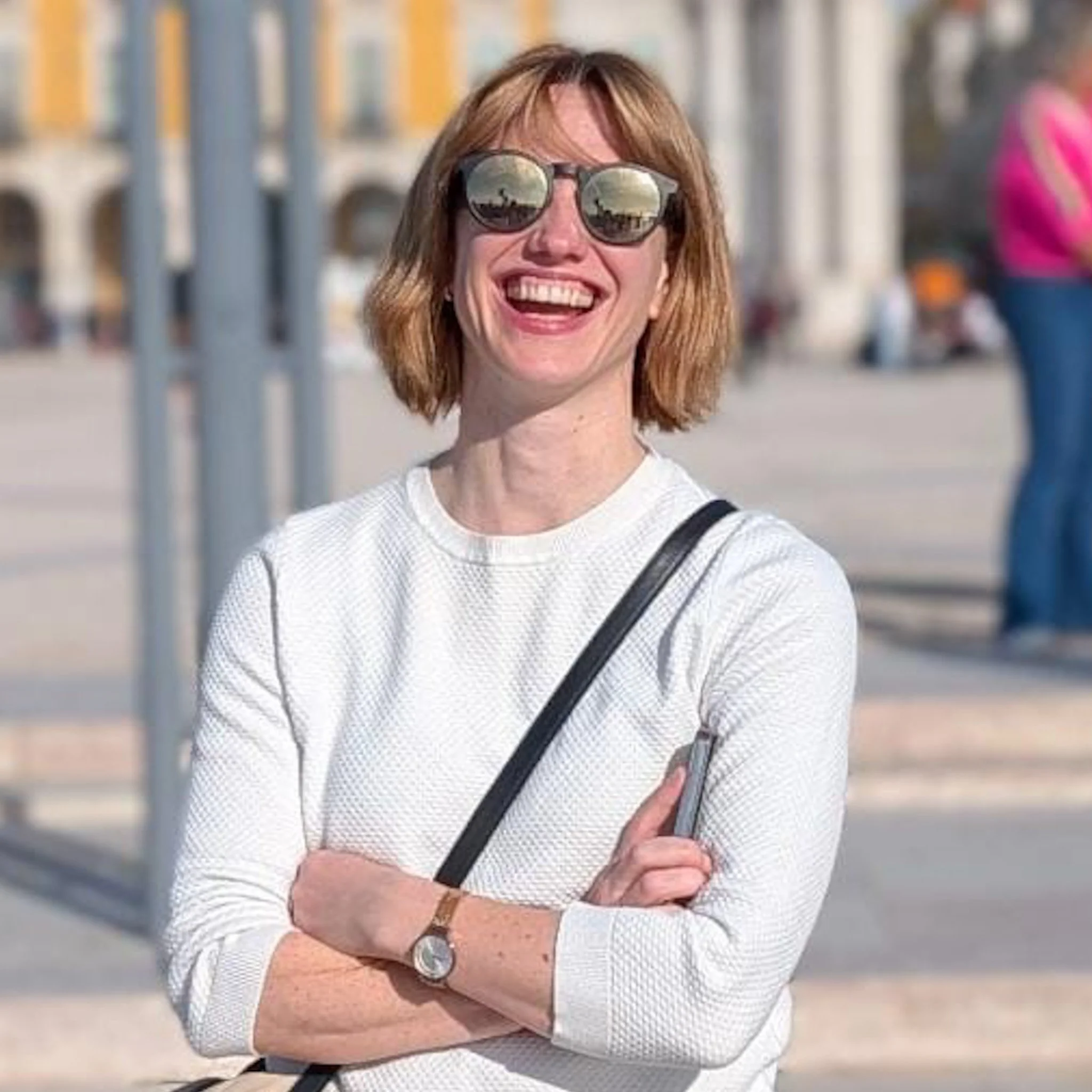 Eine lachende Frau mit roten Haaren, Sonnenbrille und weißem Oberteil steht an einer Uferpromenade, hinter ihr sind Brücken und Wasser sichtbar.