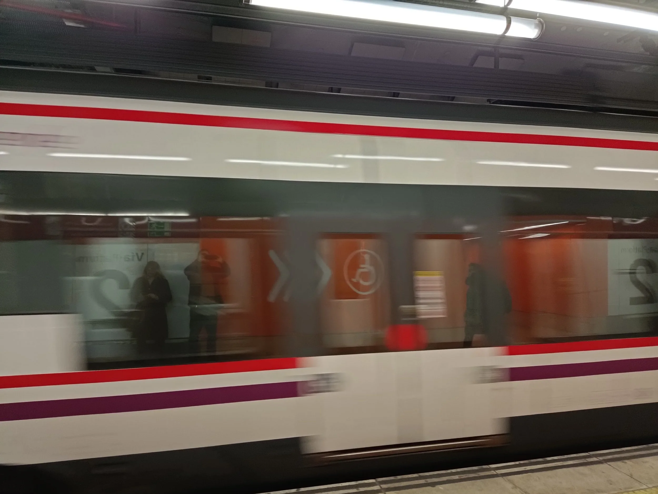 A moving train with red and purple stripes, view through a window showing people standing on the platform, and an orange wall with signs, including accessibility and directional arrows.