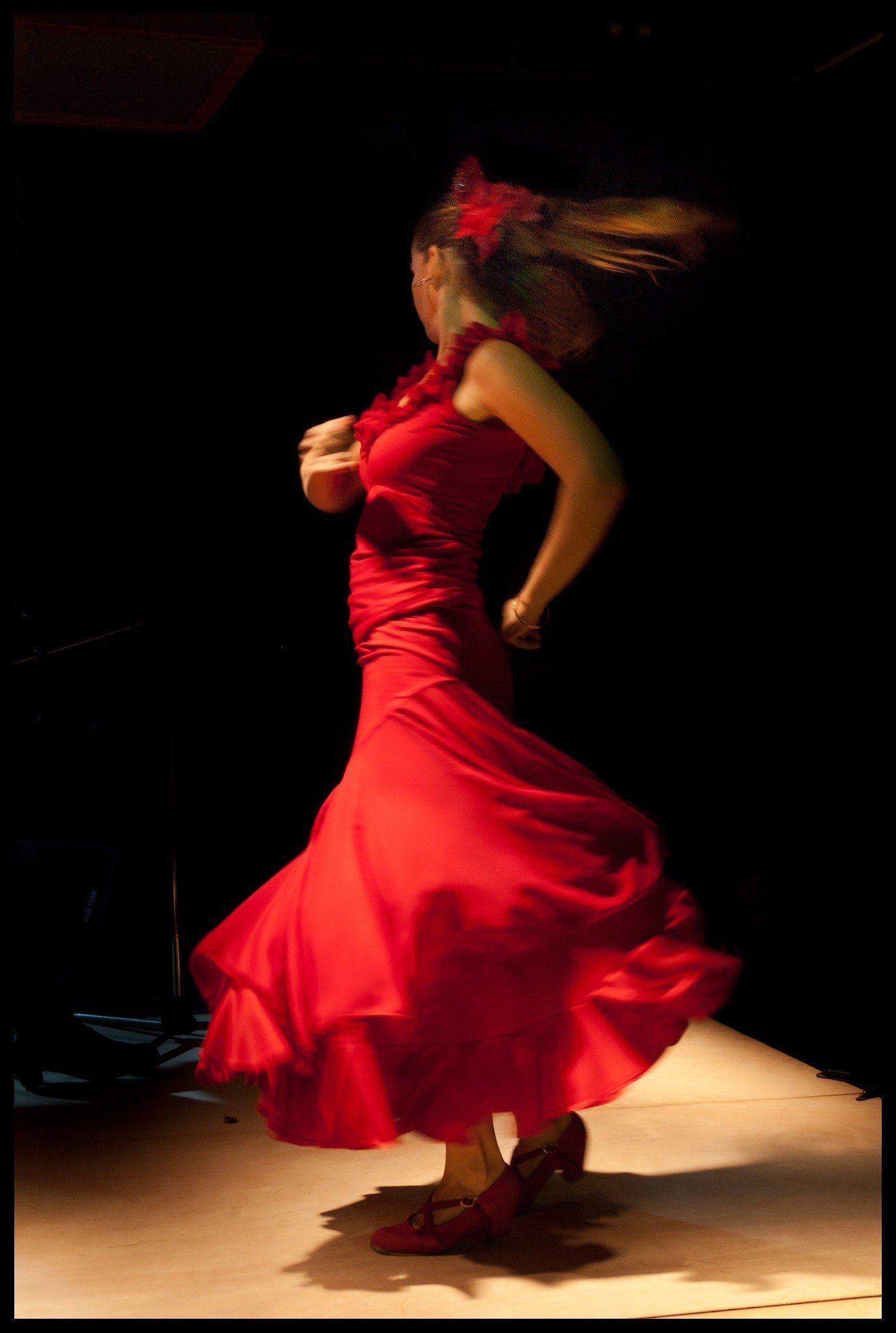 A woman dancing in a flowing red dress with ruffles, wearing red shoes, with her hair moving and a black background.