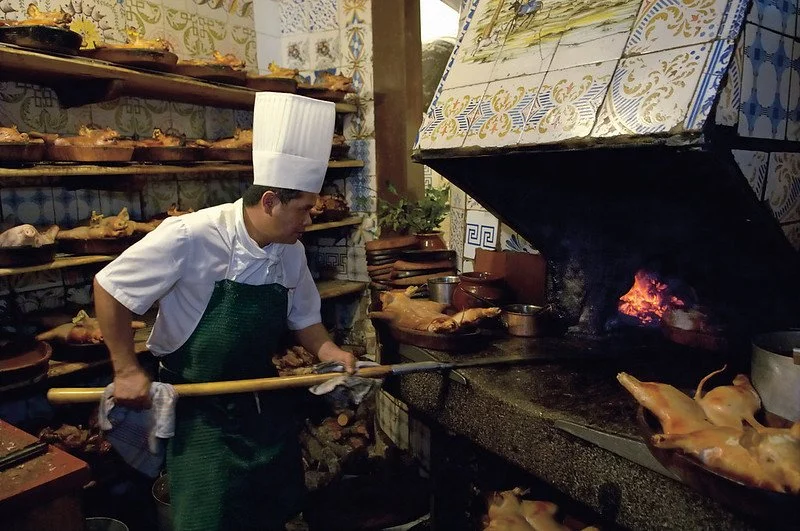 Chef roasting pigs over an open wood fire in a kitchen with traditional tile walls.