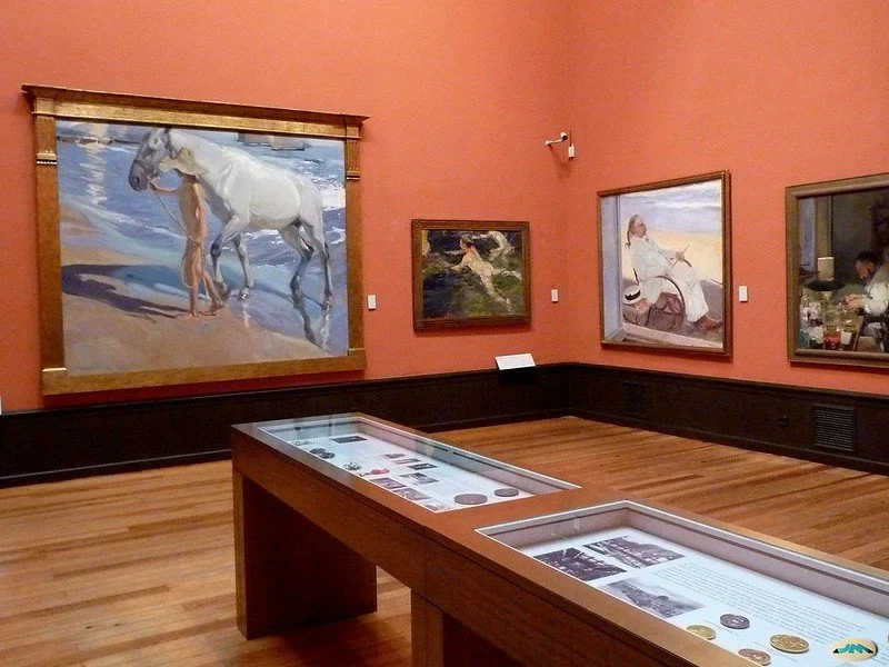 An art gallery with framed paintings on a pink wall, including a large painting of a horse and a small painting of a woman swimming. There is a display case with information and artifacts in the foreground.