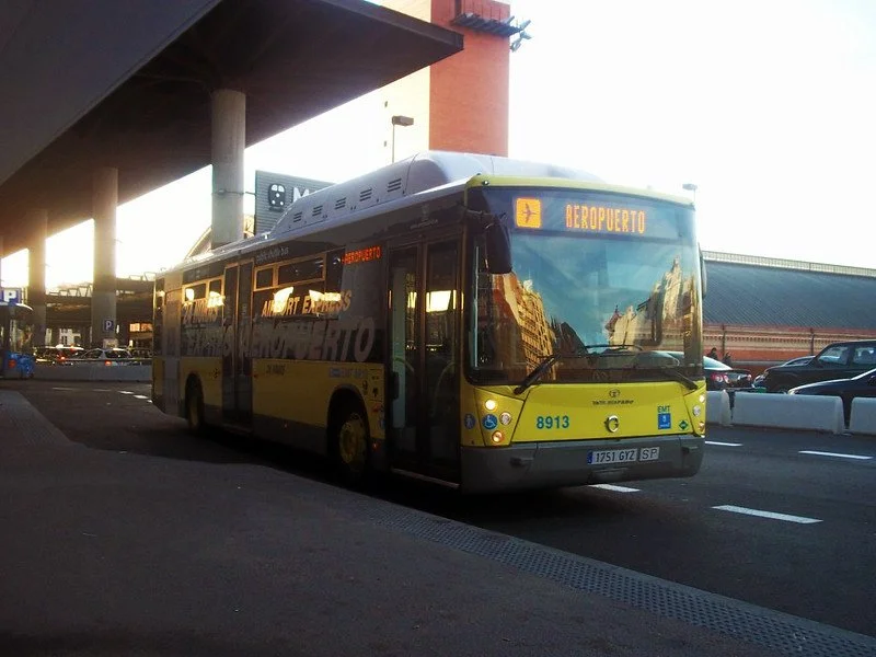 A yellow and gray public bus at an airport terminal with a digital sign indicating it is headed to the airport.