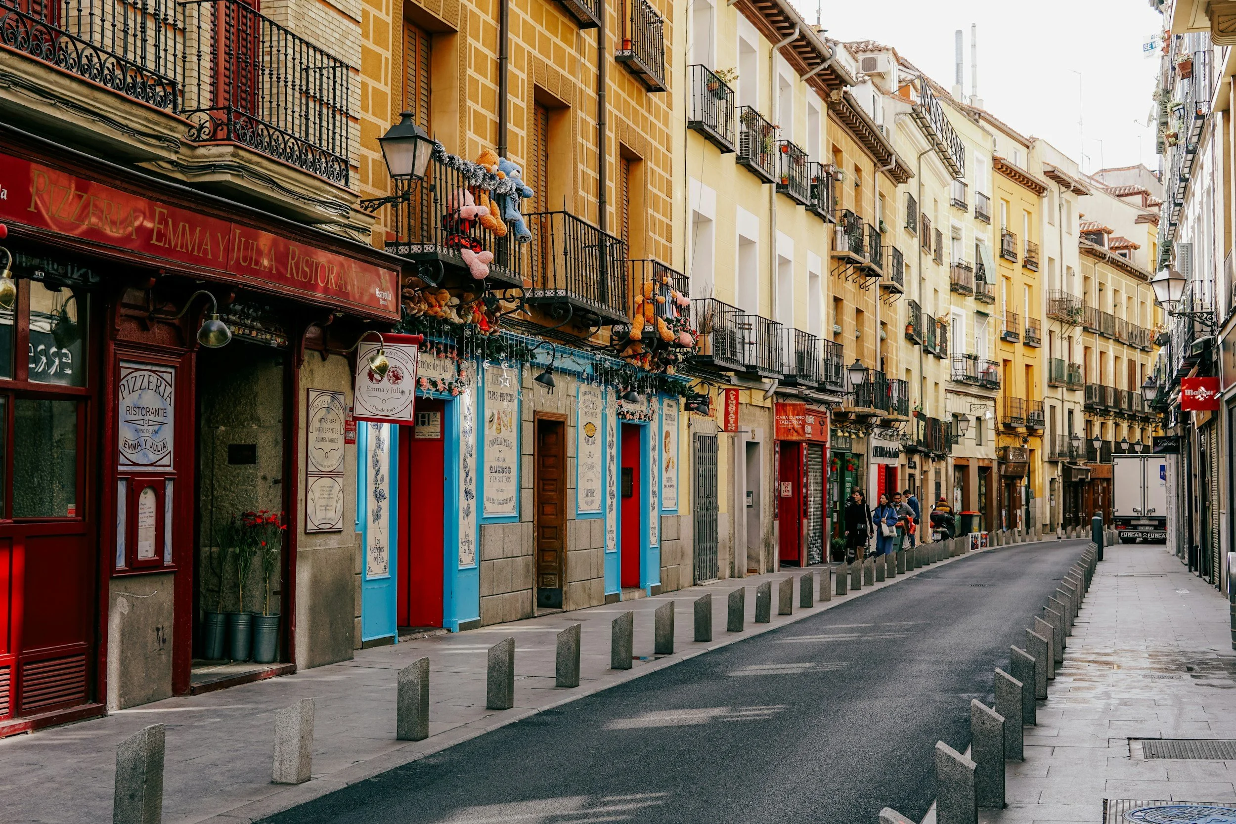 A European city street with colorful buildings, shops, and cafes. There are small balconies with plants and teddy bears, and a few people walking.