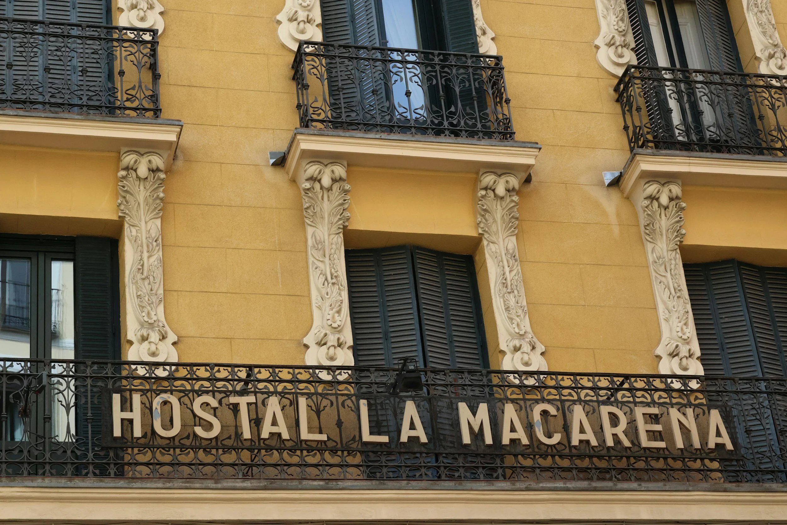 Facade of a yellow building with decorative white columns and black wrought iron balconies. A sign with black letters on a black background reads "Hostal La Macarena".