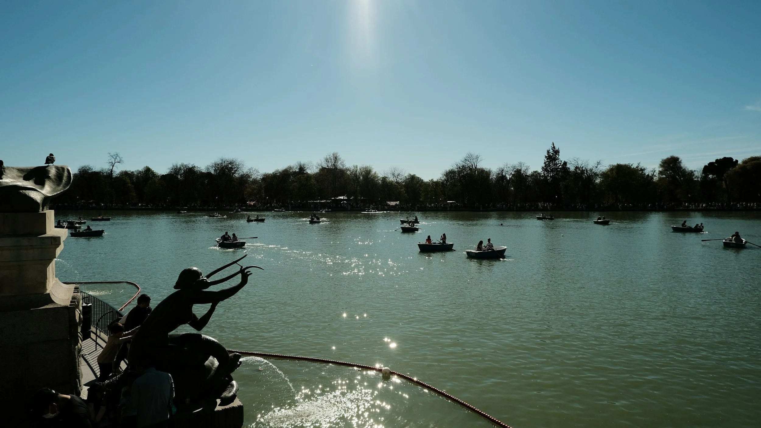 A lake with several small boats and people on the water under a clear sky, with trees on the far shore and a sculpture on the left side of the image.