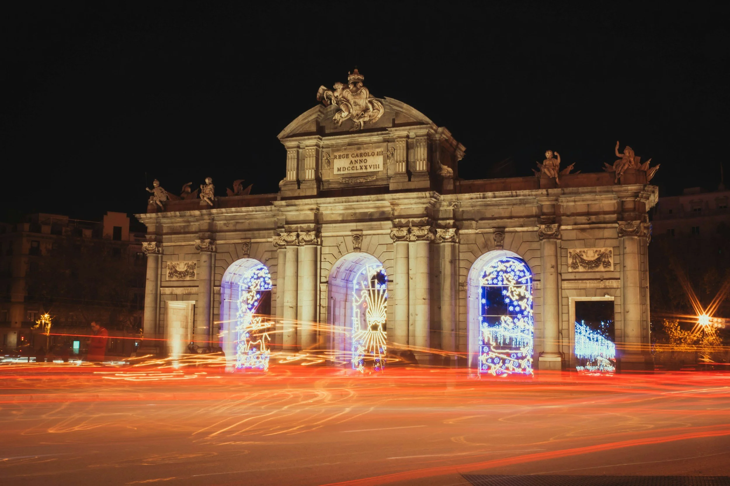 Night view of a historic archway decorated with colorful Christmas lights, with light trails from moving vehicles in the foreground.