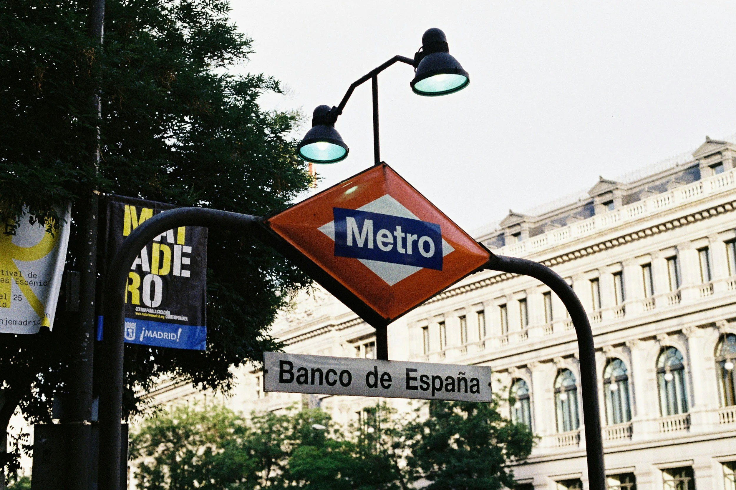 A protest sign at a Madrid metro station with a historic building and green trees in the background.