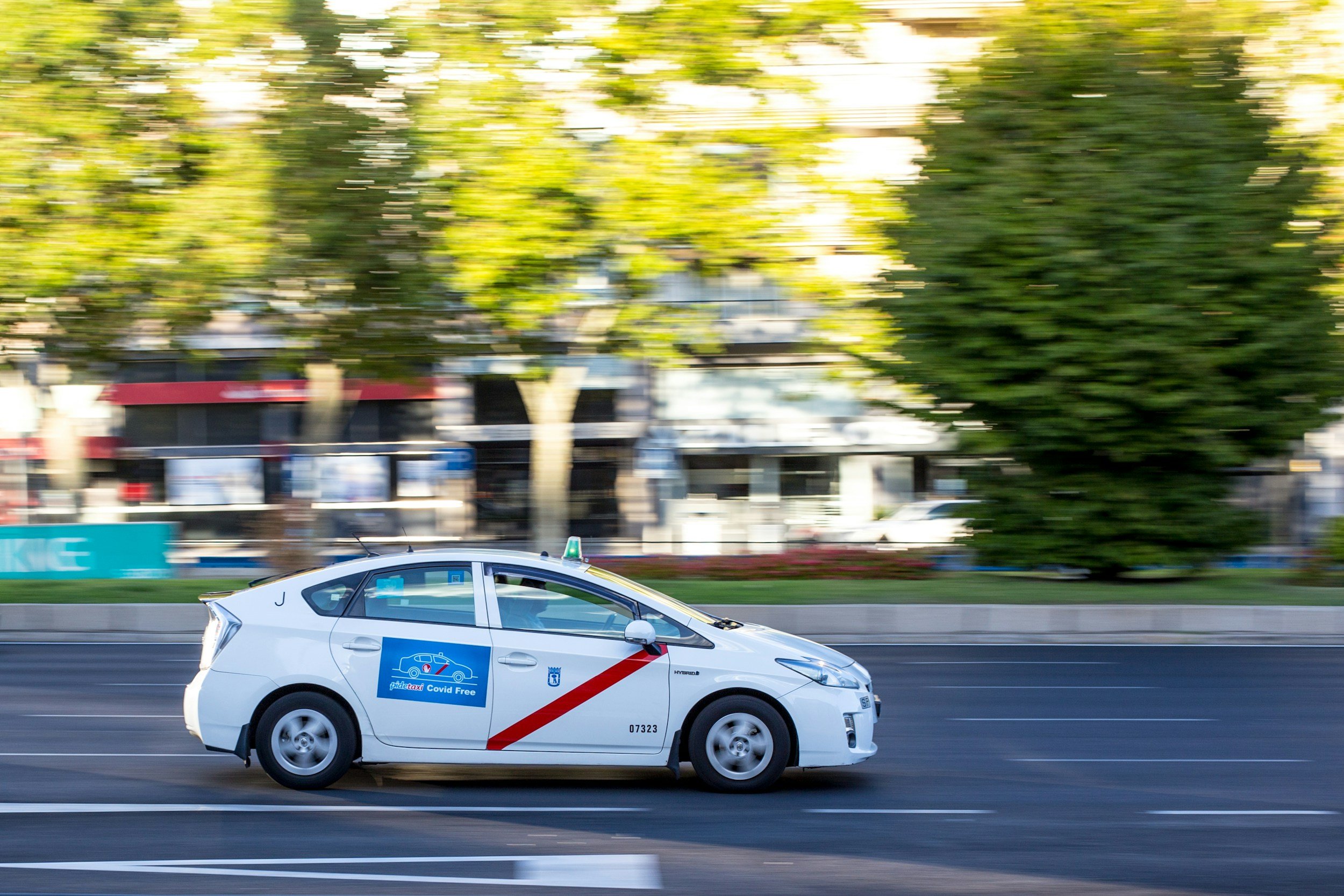 A white taxi cab driving on a city street with motion blur, trees and buildings in the background.