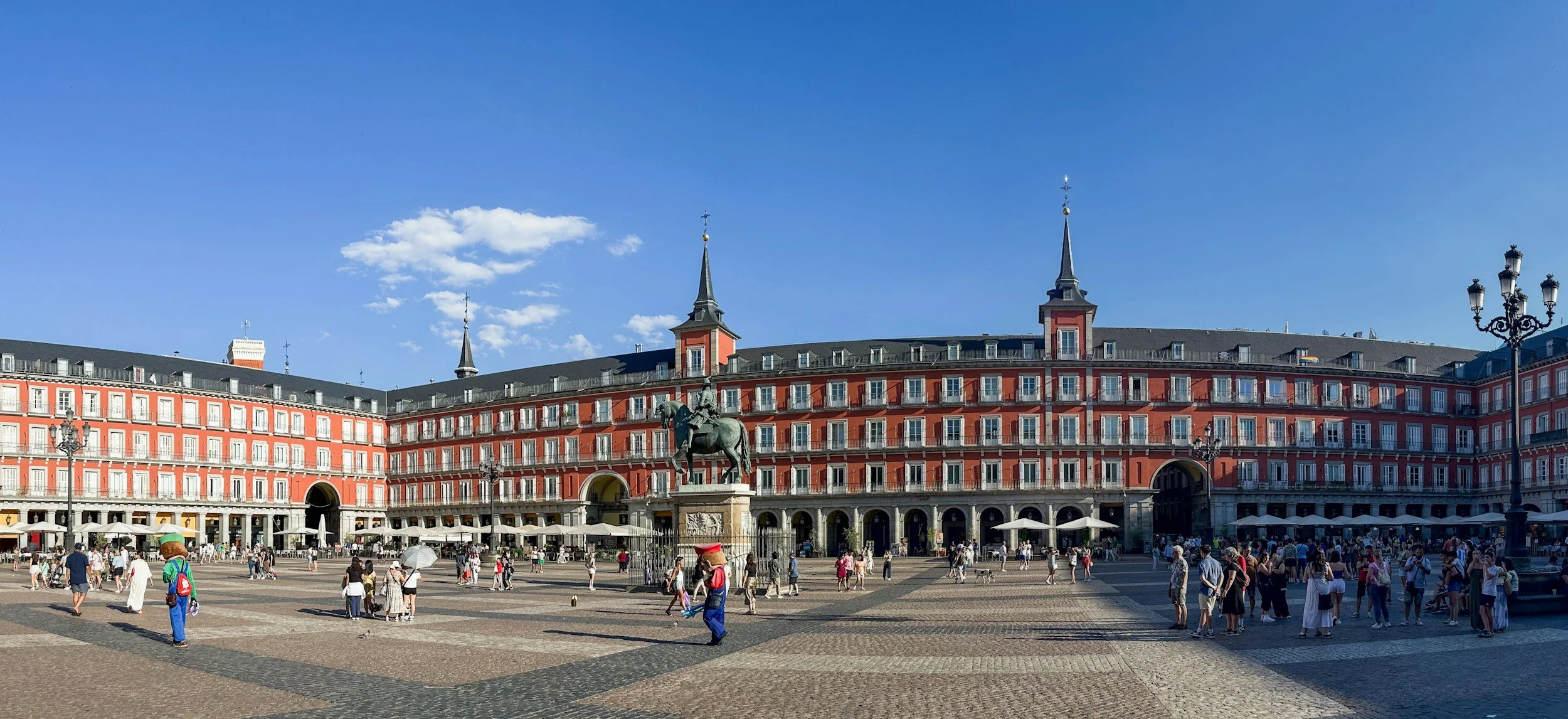 Public square with a red building, statue of a man on horse, crowds walking, umbrellas, and streetlights under a blue sky.