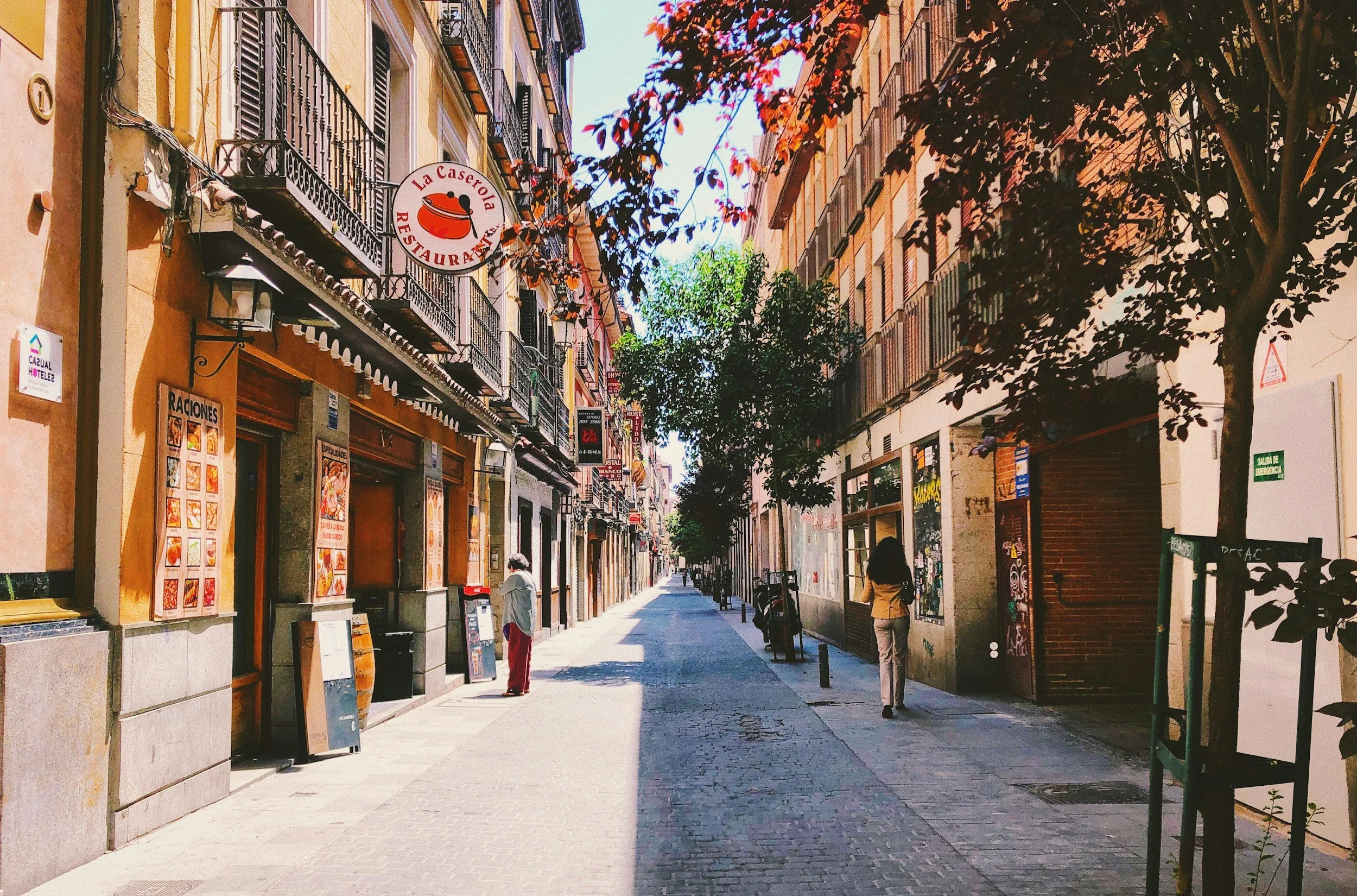 A quiet city street with shops and restaurants on both sides, trees lining the sidewalk, and a woman walking away from the camera in the distance.
