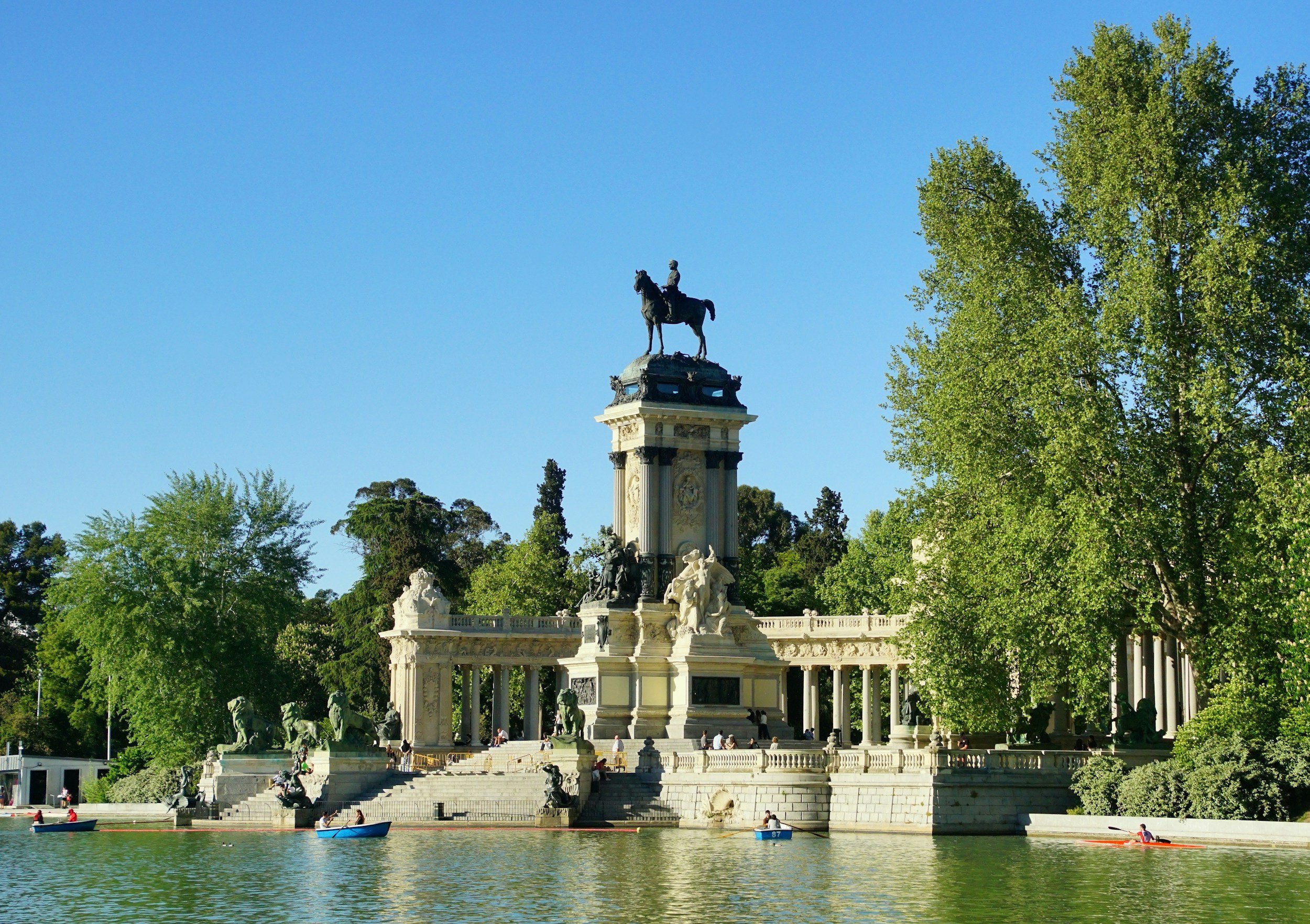 Historical monument with a large statue of a person on a horse at the top, surrounded by sculptures and columns, with a water body in the foreground and trees in the background.