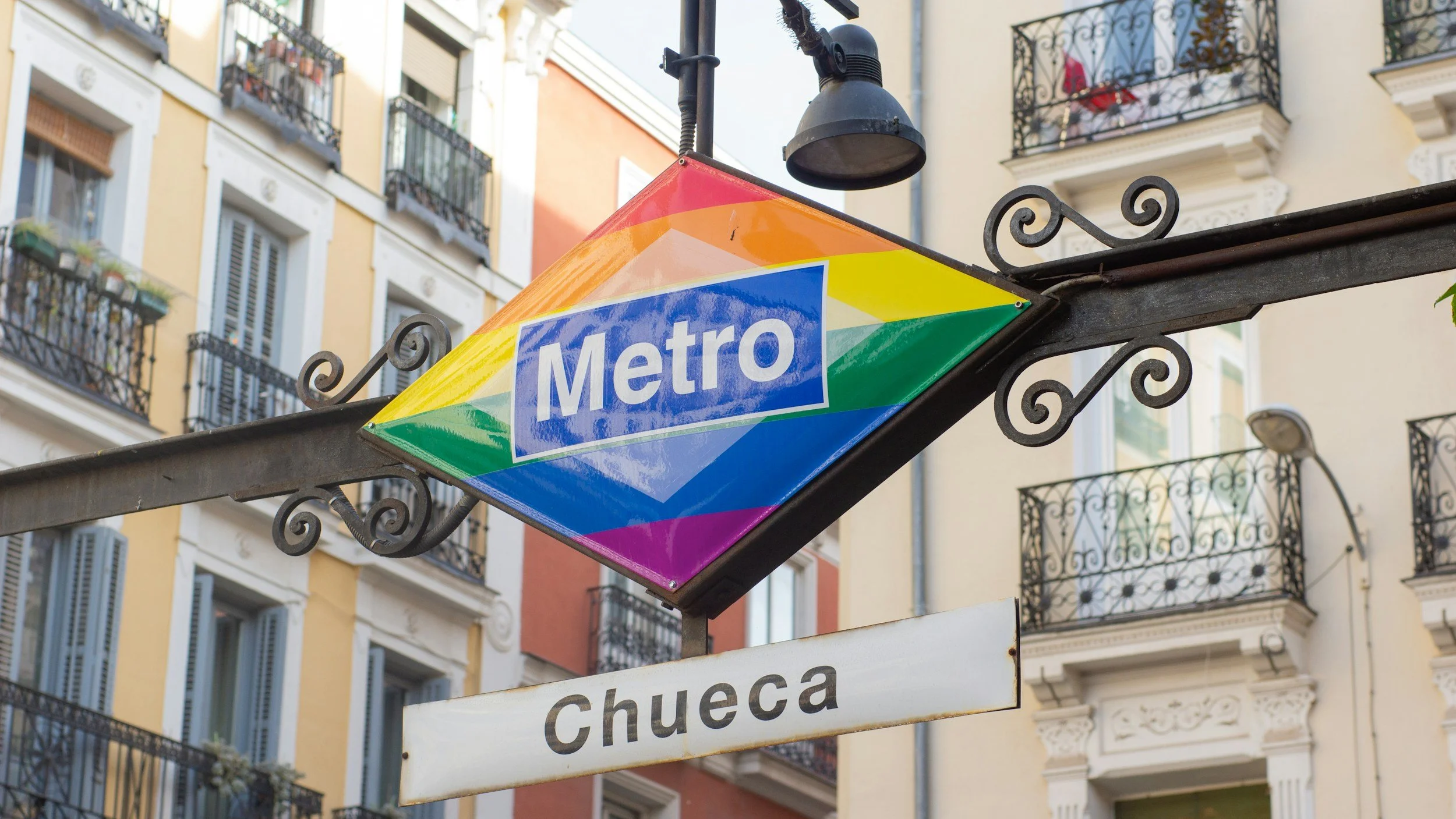 Colorful Metro sign with rainbow stripes, hanging in front of European-style apartment buildings with balconies.
